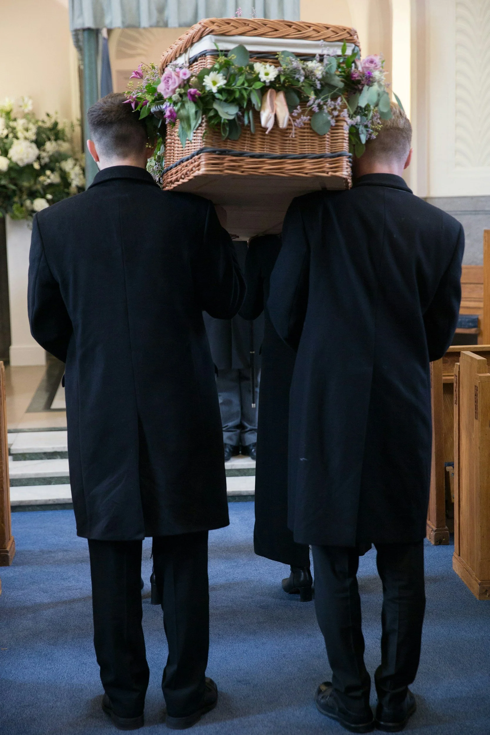 Two individuals dressed in black suits carry a wooden casket decorated with flowers during a thomas brothers funeral service.