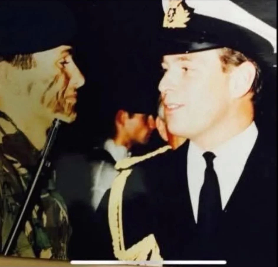 A young man in a police uniform smiling and talking to a woman in a hat, with other people in the background.