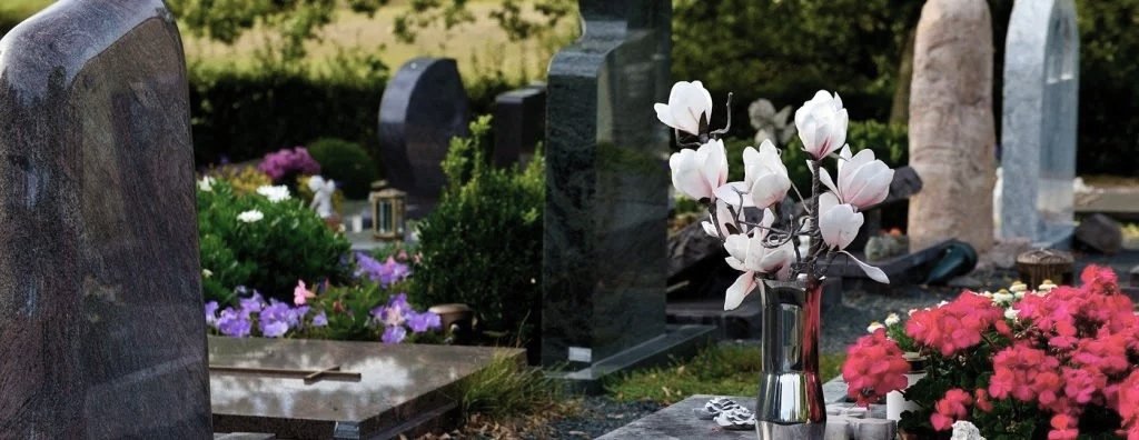 Gravestone with a bouquet of white magnolia flowers in a silver vase, surrounded by colorful flowers and other gravestones in a cemetery.