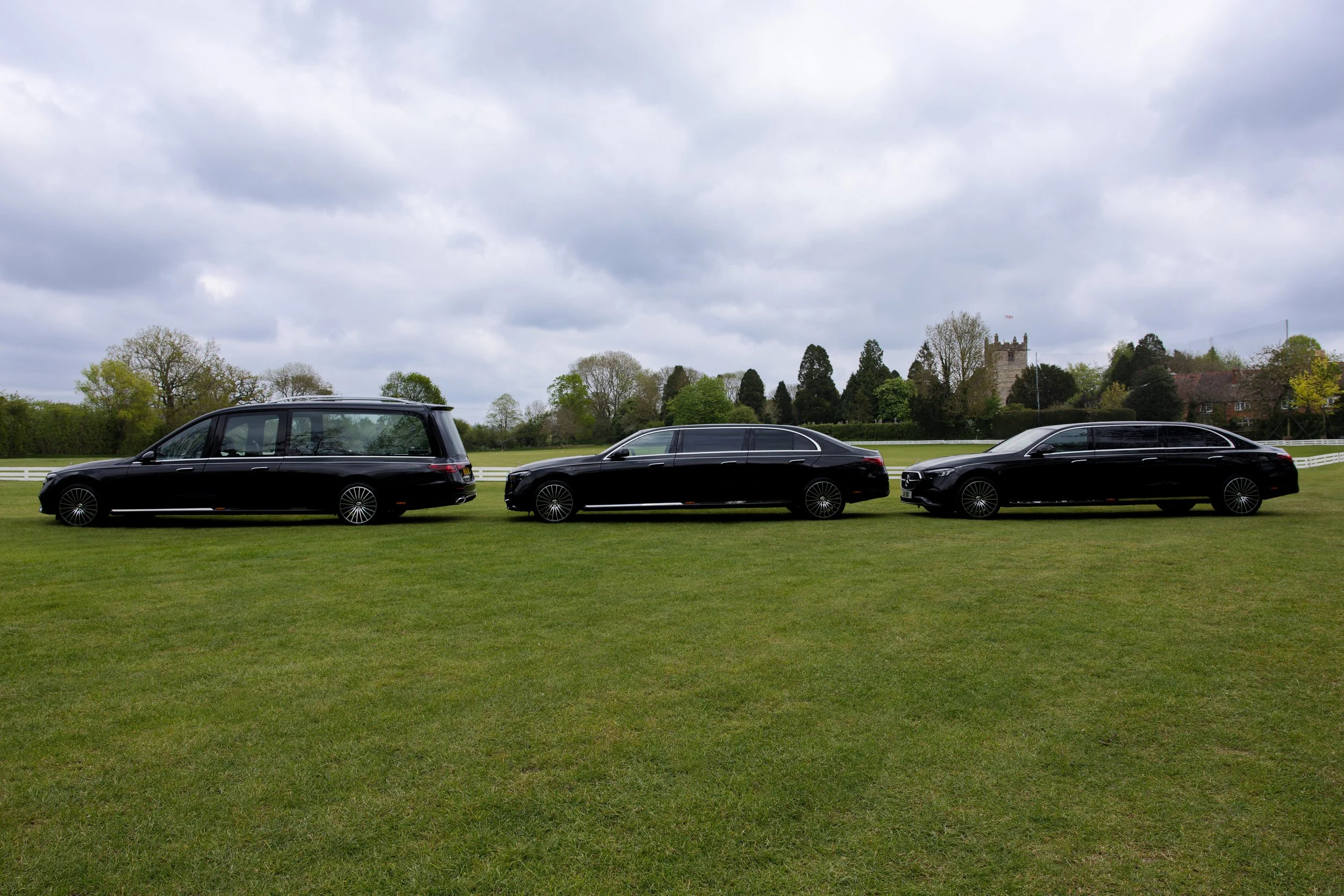 Three black luxury cars parked on a grassy field with trees in the background, a castle-like building on the right, and a cloudy sky overhead.