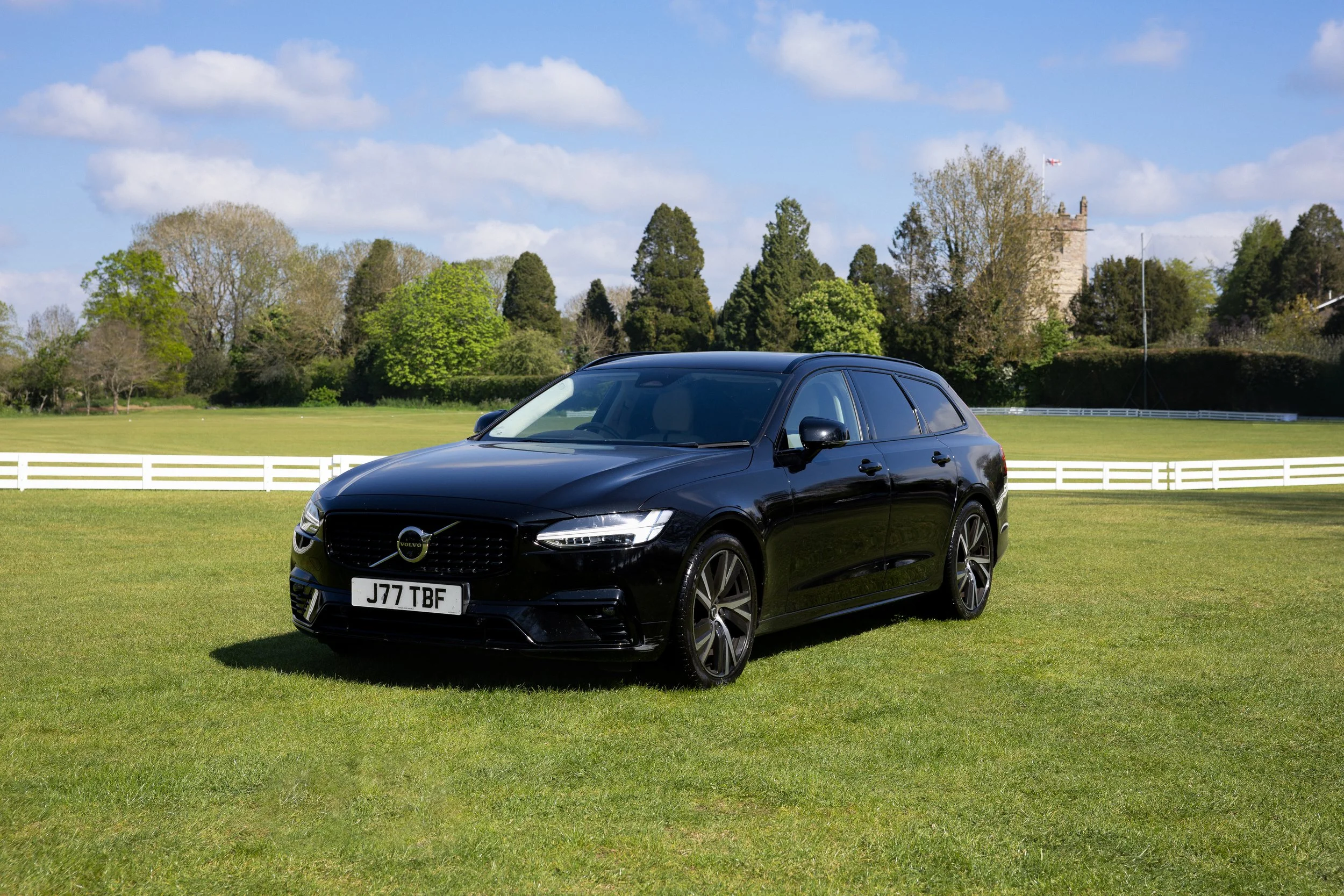 A black Volvo SUV parked on a grassy field with trees and a castle in the background under a partly cloudy sky.