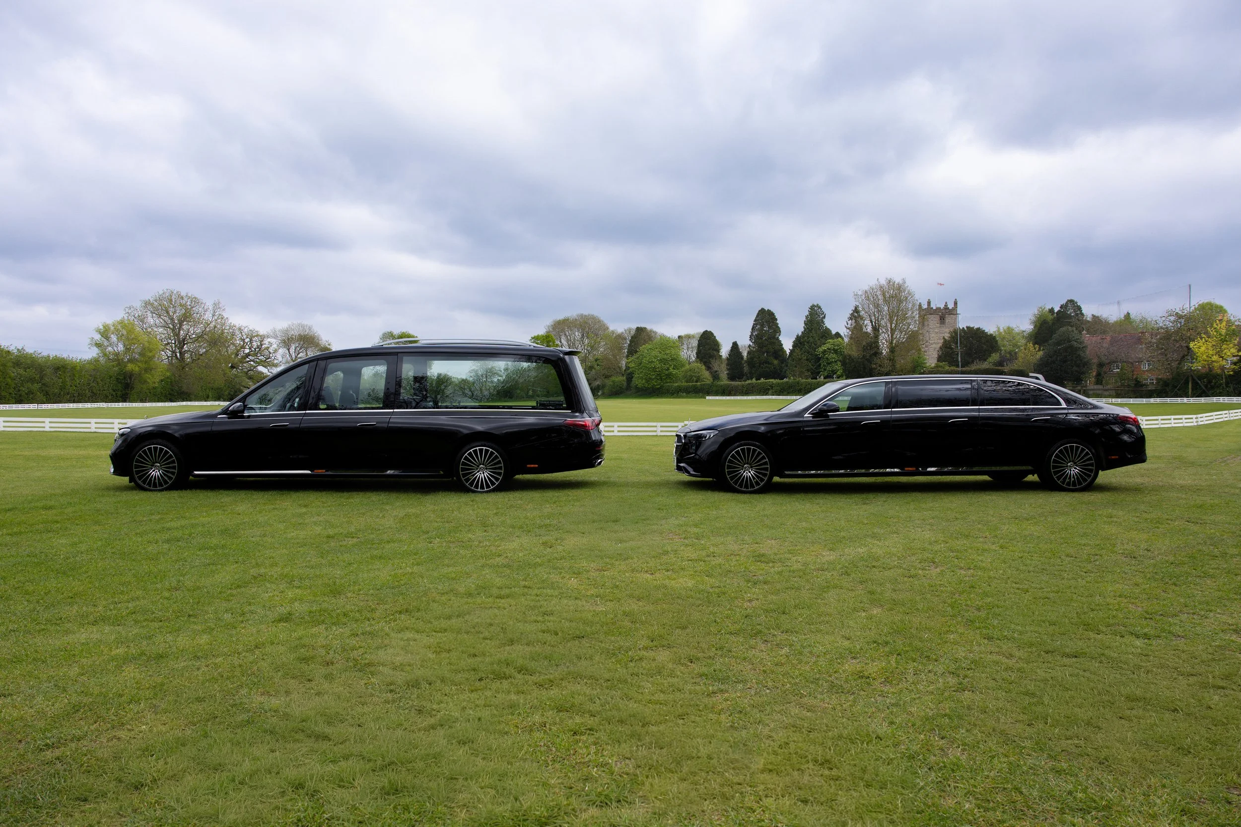 Two black luxury cars parked on a grassy field with a white fence, trees, and a stone castle in the background under a cloudy sky.