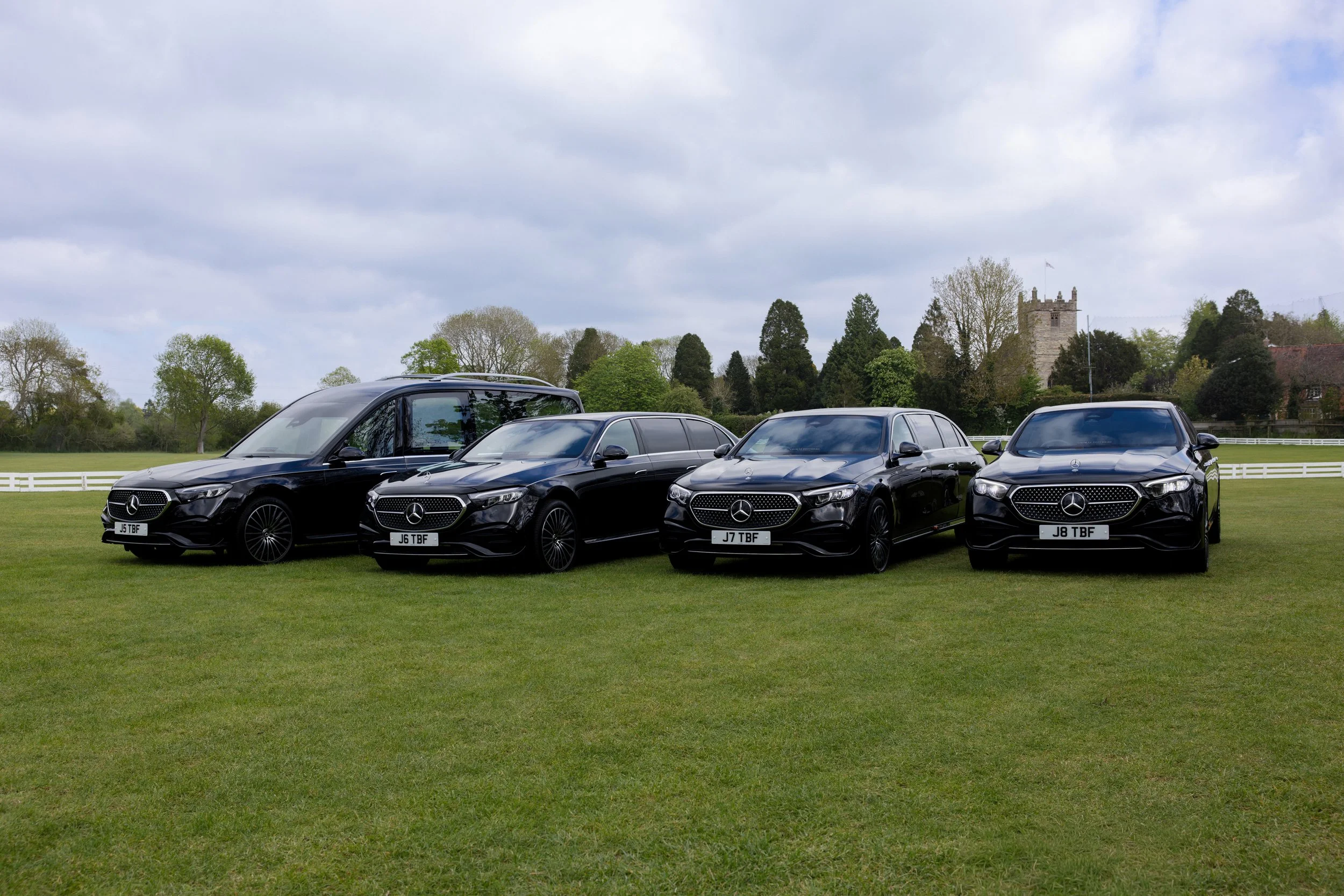 Four black Mercedes-Benz cars parked on a grassy field with trees and a church tower in the background.