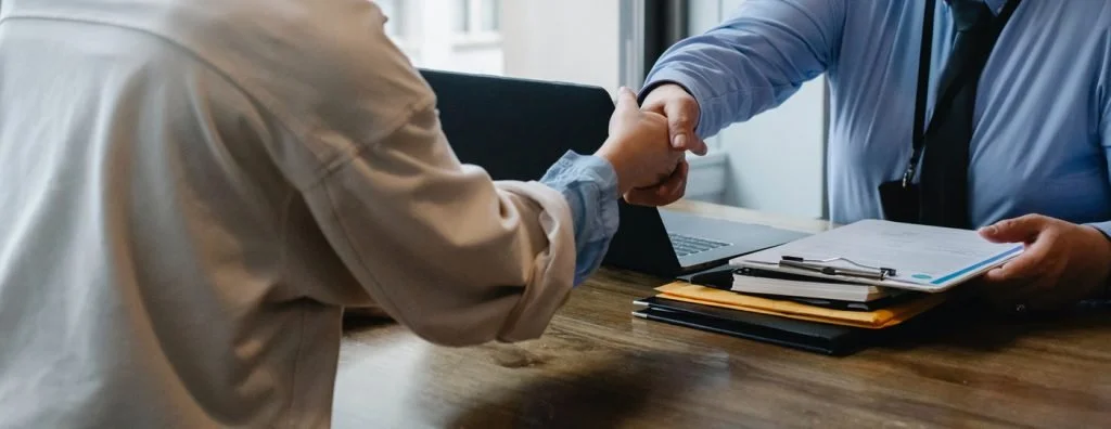 Two people shaking hands across a desk in an office setting, with documents and a laptop present.