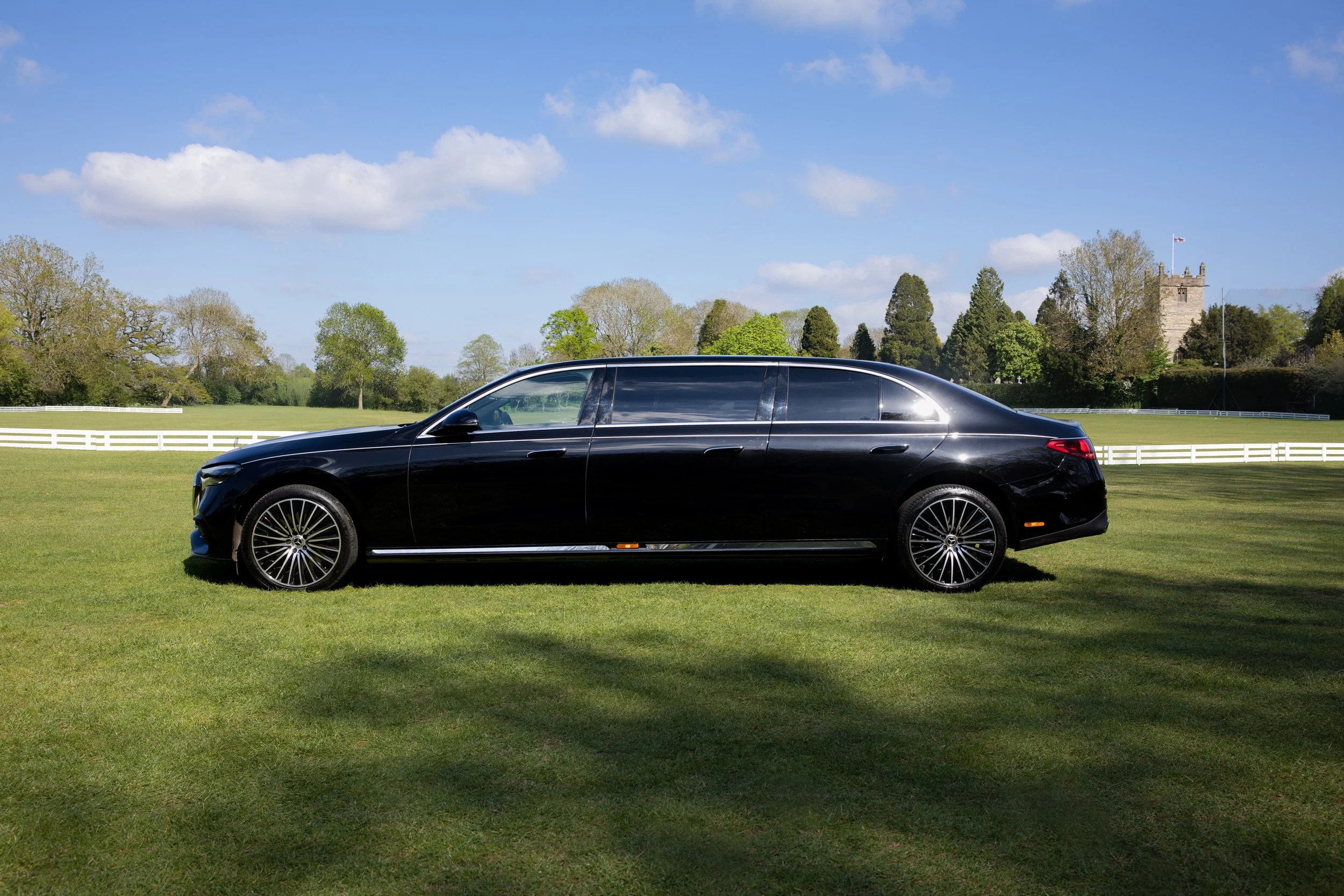 A sleek black luxury sedan parked on a wide grassy field with trees, a tower, and a fence in the background under a blue sky with clouds.