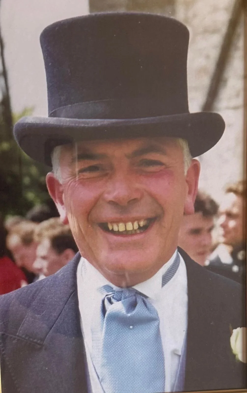 A man with a big smile wearing a top hat, suit, and tie, at an outdoor event with people in the background.