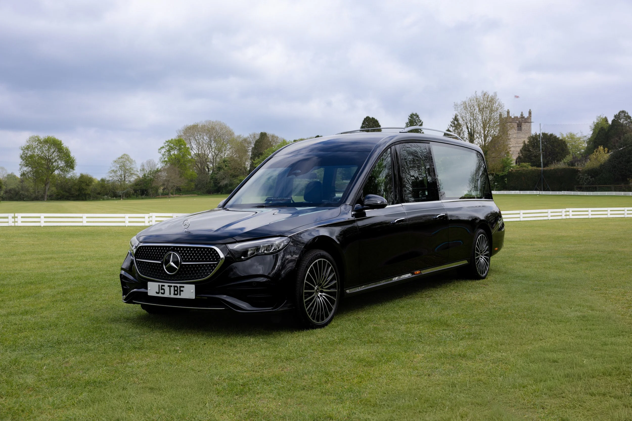 A black Mercedes-Benz car parked on a green field with trees and a castle in the background under cloudy skies.