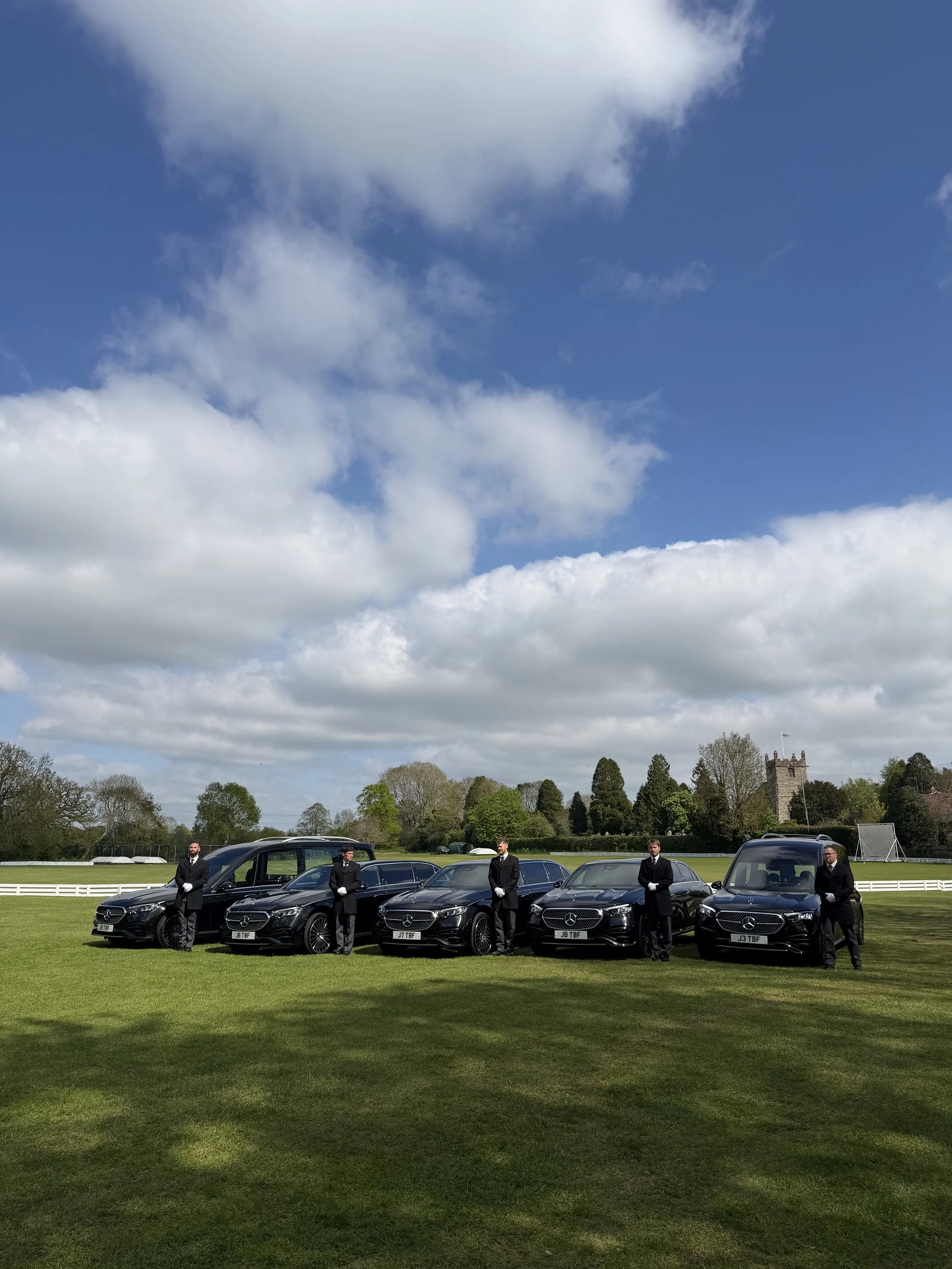 Five men in black suits and white gloves standing next to luxury black Mercedes-Benz cars on a green field, with trees, a tower, and a cloudy sky in the background.