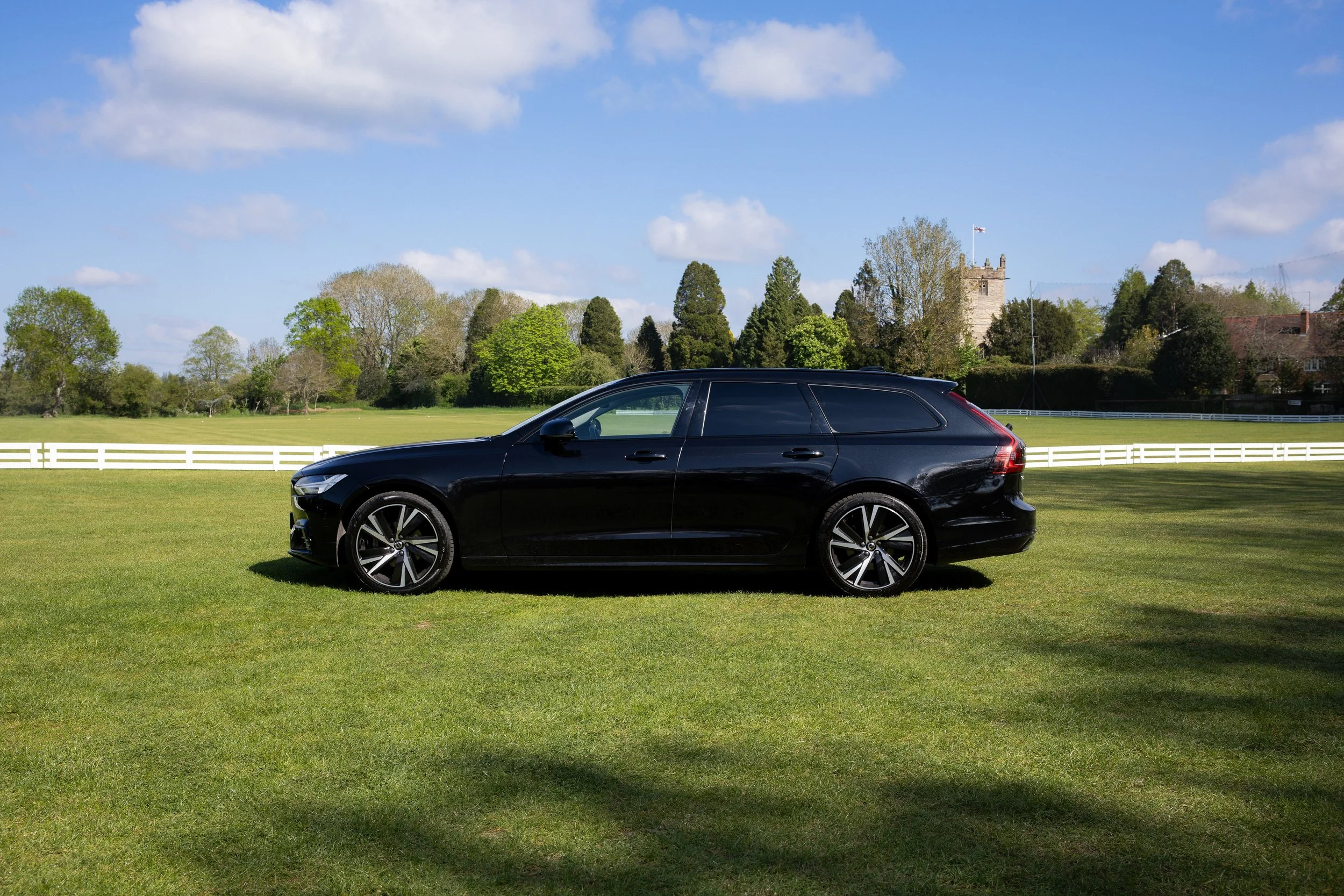 Black car parked on a grassy field with trees and a castle in the background under a partly cloudy sky.
