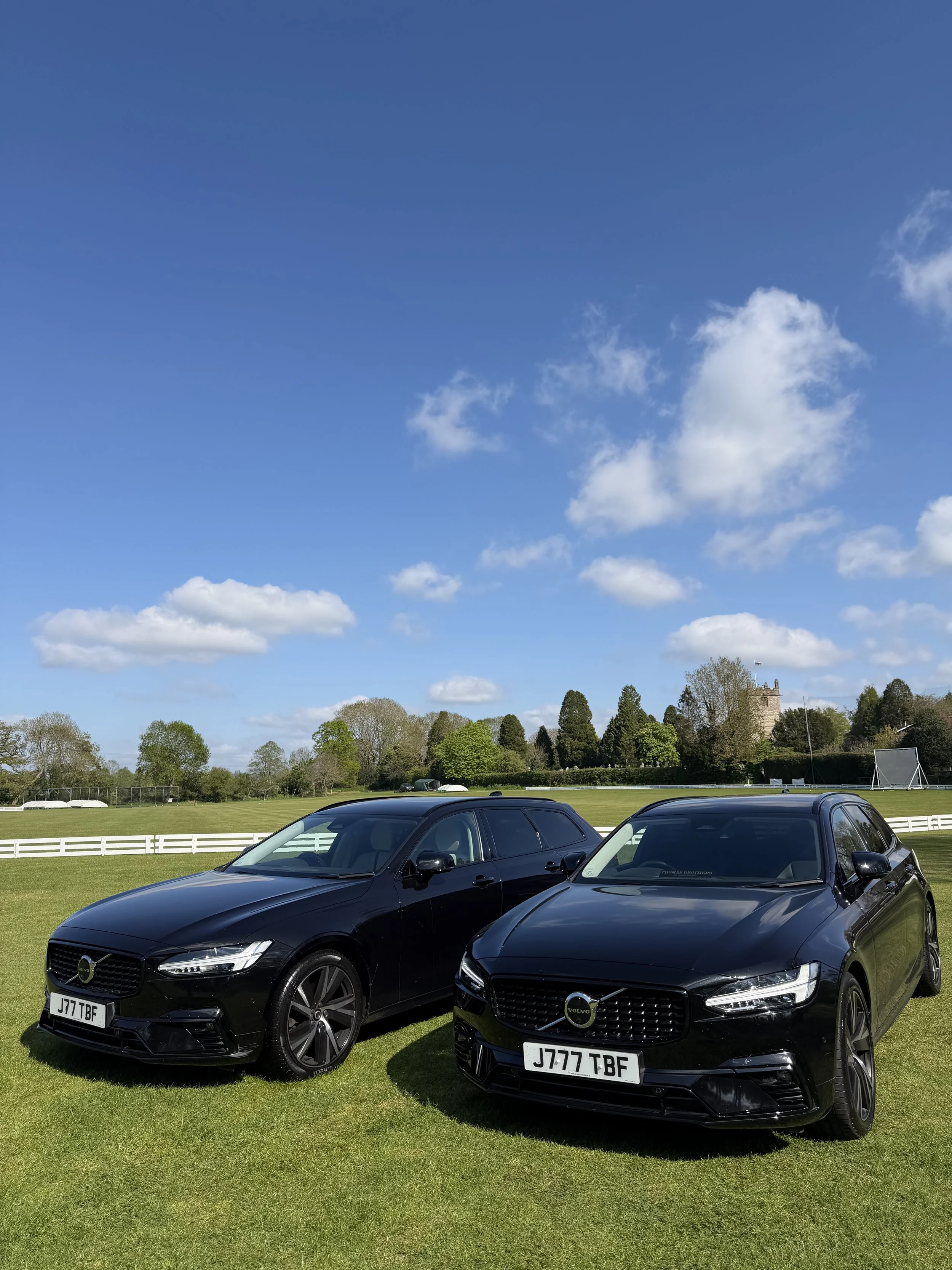 Four black cars parked on a grassy field with a white fence, trees, a castle-like building in the background, and a bright blue sky with some white clouds.