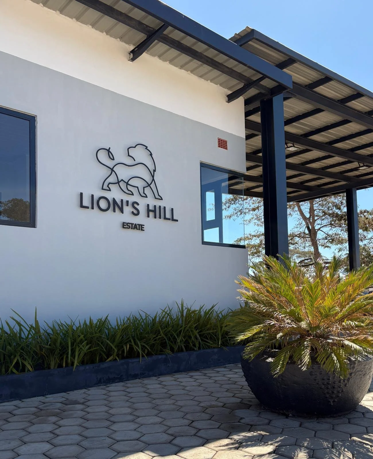 Exterior of Lion's Hill Estate Gatehouse with a sign that reads 'Lion's Hill Estate' accompanied by a minimalist lion logo, with a large potted plant in the foreground and clear blue sky overhead.