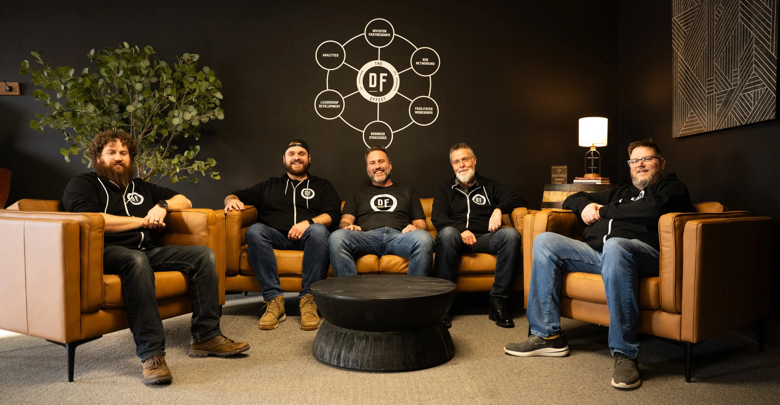 Five men sitting on tan leather sofas in a room with black walls, smiling and wearing black hoodies with white logos, with a large green plant on the left and a round black coffee table in front. Behind them is a wall graphic titled 'The DF Effect' with various connected terms.