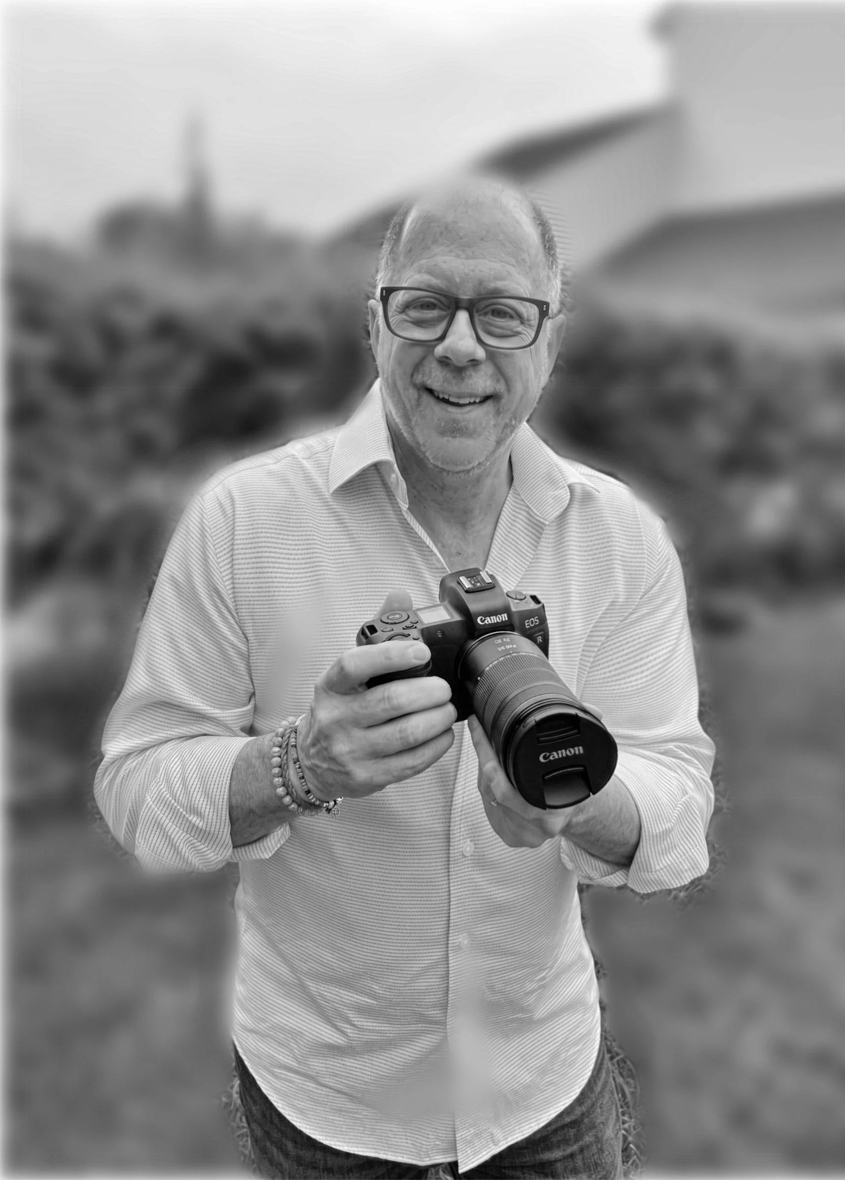 Black and white photo of a smiling man with glasses, holding a Canon EOS camera, outdoors in a blurry natural setting.