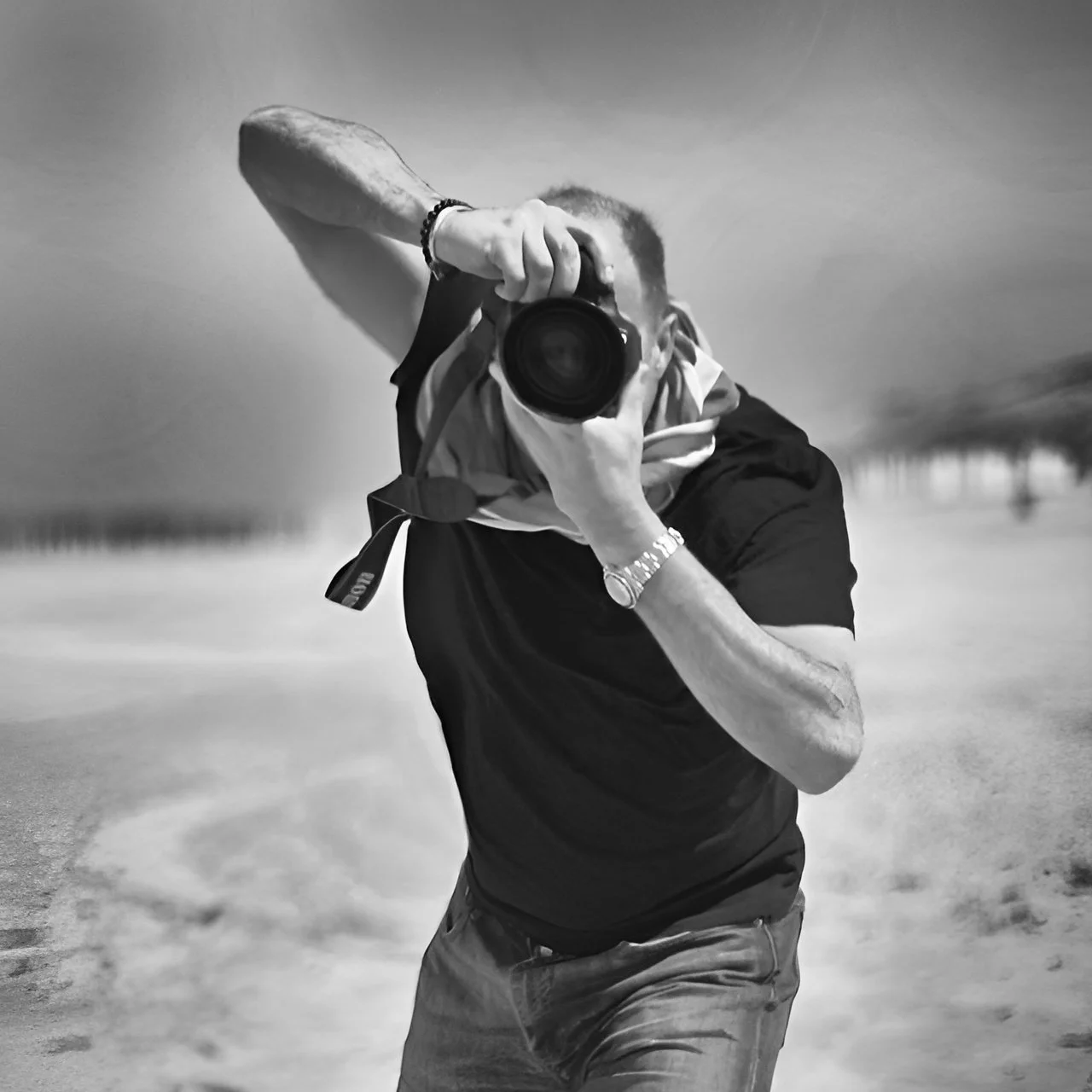 A man taking a photo with a camera outdoors, wearing a black shirt, jeans, a watch, and a scarf over his shoulder, with a blurred landscape background.