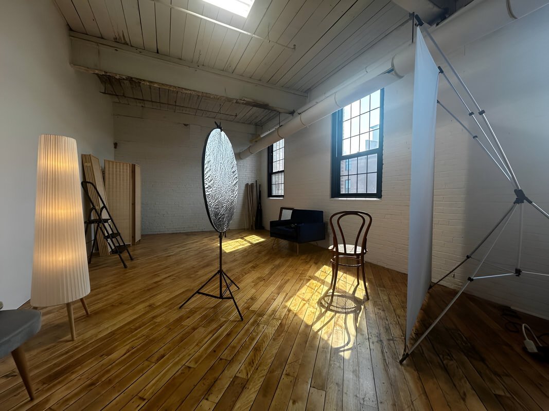Interior of a photography studio with wooden floors, white painted brick walls, large windows, a studio light reflector, a tripod with a white backdrop, a floor lamp, and some chairs.