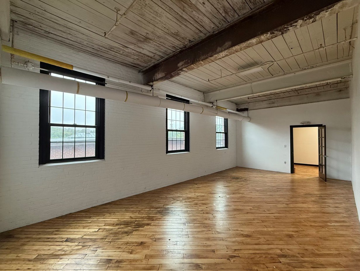 Empty loft space with three large windows, wooden floors, exposed wooden ceiling, and a door leading to another room.