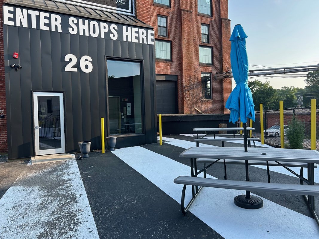 Urban outdoor shopping area entrance with a black corrugated metal wall, large sign saying 'ENTER SHOPS HERE,' white numbers '26,' a door, and outdoor tables with a blue umbrella, yellow safety posts, benches, and a black pavement with a painted white stripe.