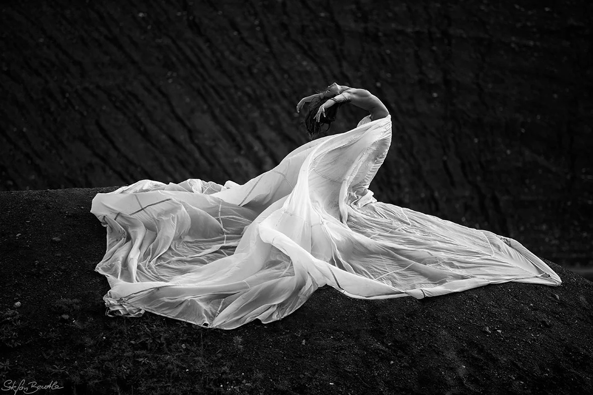 A woman in a flowing white dress poses on dark, textured ground with her head thrown back and arms raised above her head.
