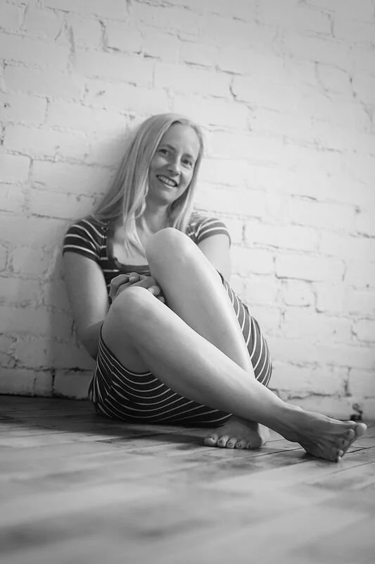 A woman with long blonde hair sitting on the floor against a white brick wall, smiling, wearing a striped dress.
