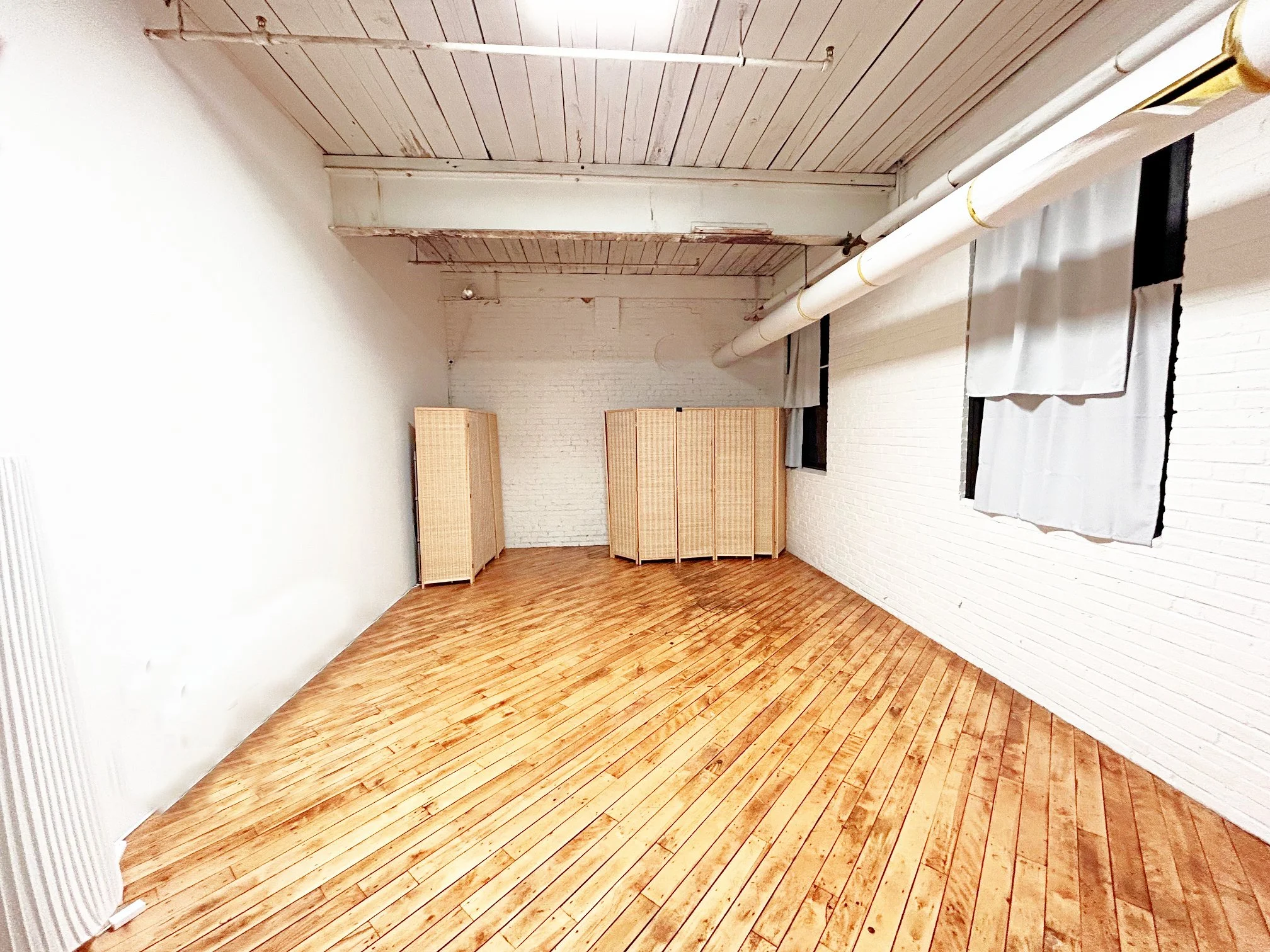An empty room with white brick walls, a wooden floor, a white ceiling with visible pipes, and a three-panel folding screen against the far wall. There are windows on the right side with white curtains.