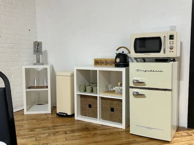 Kitchen area with a white refrigerator, microwave on top, white shelving unit with cups and baskets, a small white side table with a glass container, and a black kettle, on wooden flooring against a white wall.
