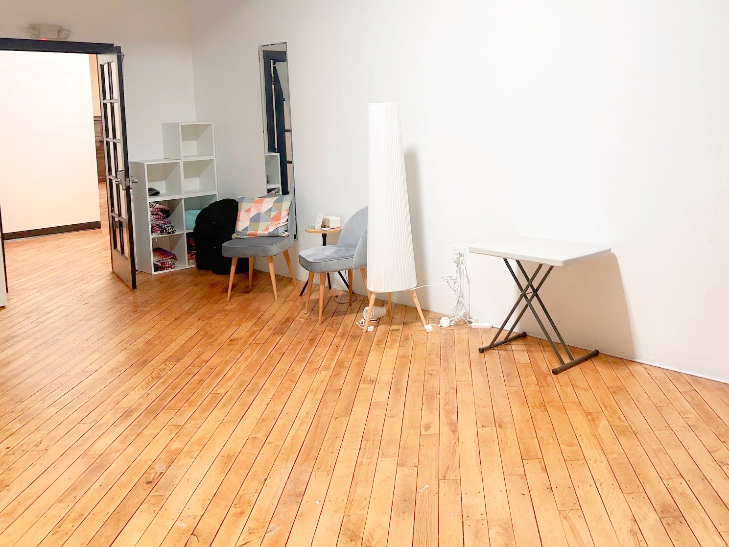 Empty room with wooden floors, white walls, a folding table, a tall white lamp, two gray chairs, and a white shelf with folded clothes near a door opening to another room.