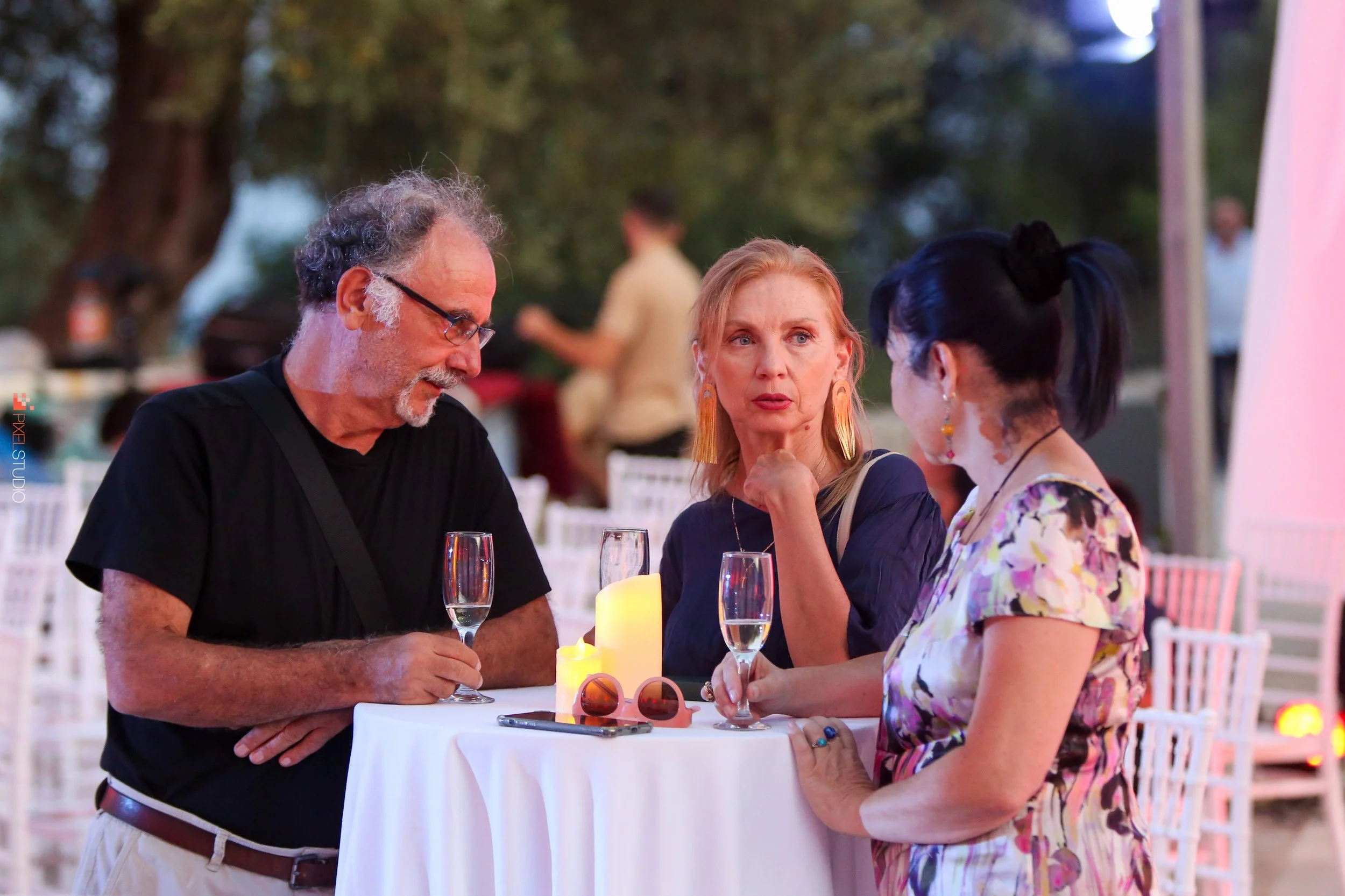 Three women are sitting at a round table with lit candles and glasses of drinks, having a conversation outdoors during the evening.
