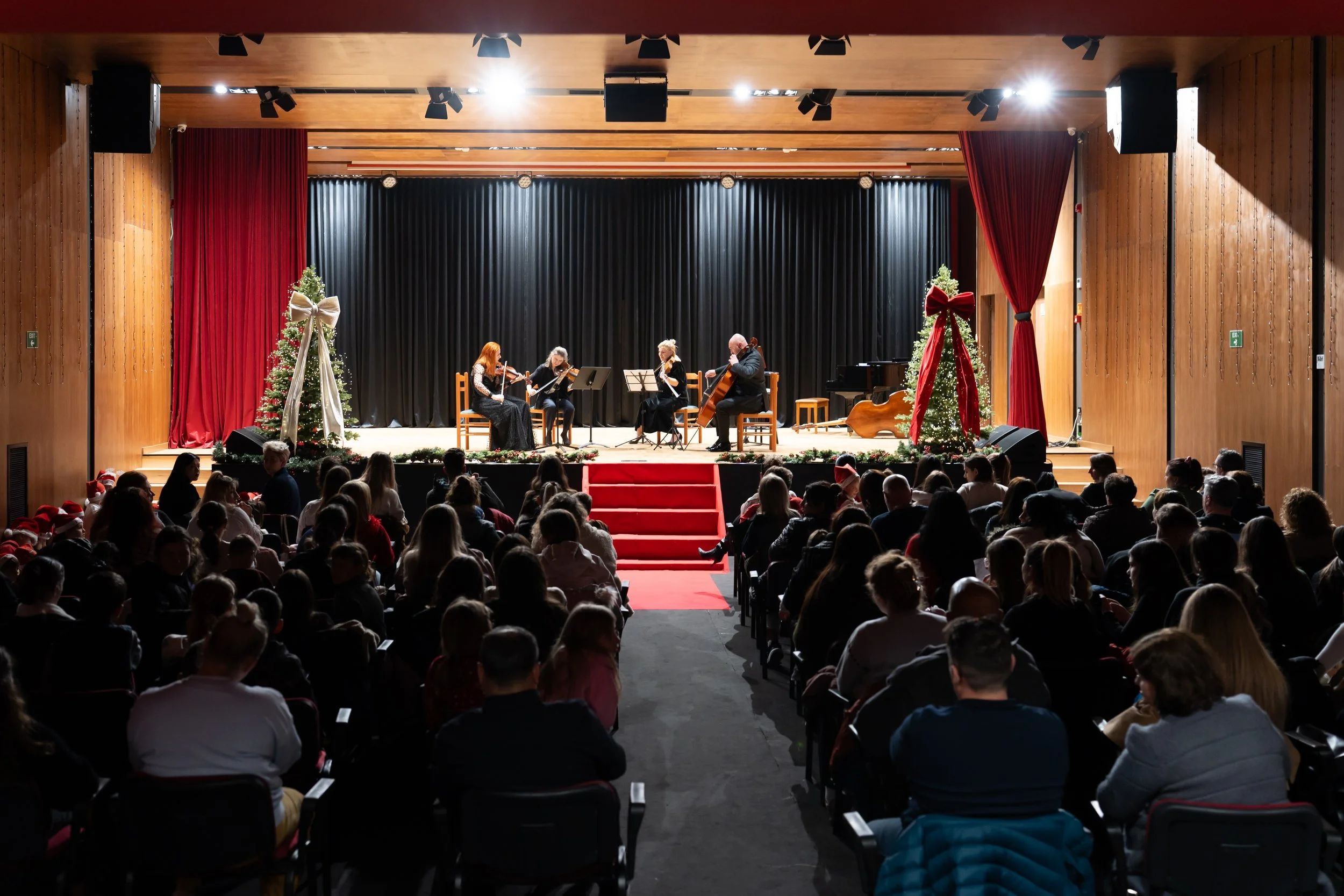 A holiday concert on a stage decorated with Christmas trees and large bows, with an audience watching.
