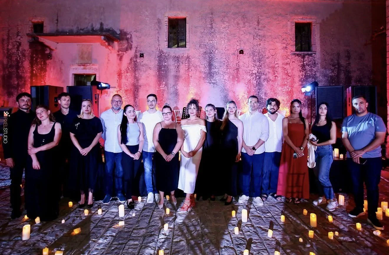 Group of 16 people standing together on a cobblestone ground with lit candles, against a textured, pink-lit wall at night.
