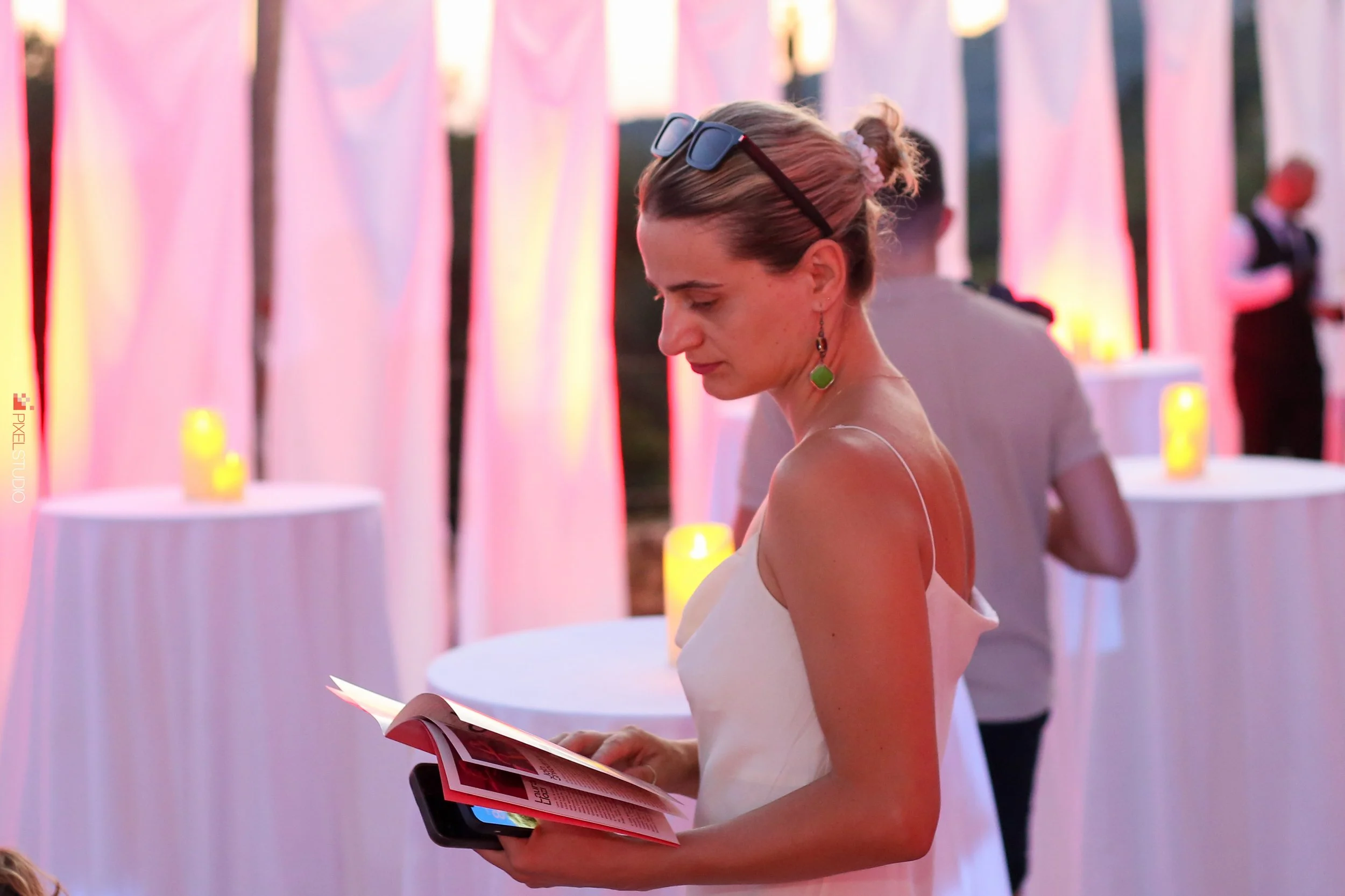 A woman with sunglasses on her head and green earrings reading a brochure at an outdoor event with pink and purple lighting.