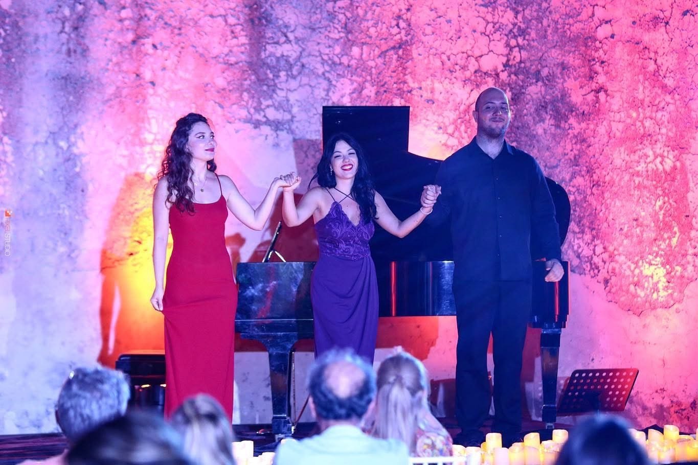 Three performers on stage, two women in elegant dresses and a man in a black shirt, holding hands and bowing at the end of a performance, with audience and candles in the foreground.