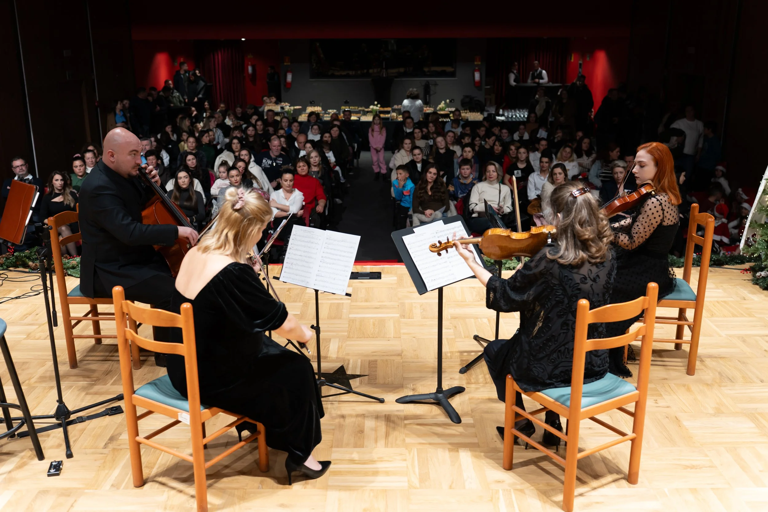 Musicians perform a concert with an audience in a dimly lit hall with red walls. The orchestra includes violinists and a cellist, viewed from behind on a wooden stage, with sheet music in front of them. The audience, composed mainly of women and children, is seated facing the musicians, watching attentively.