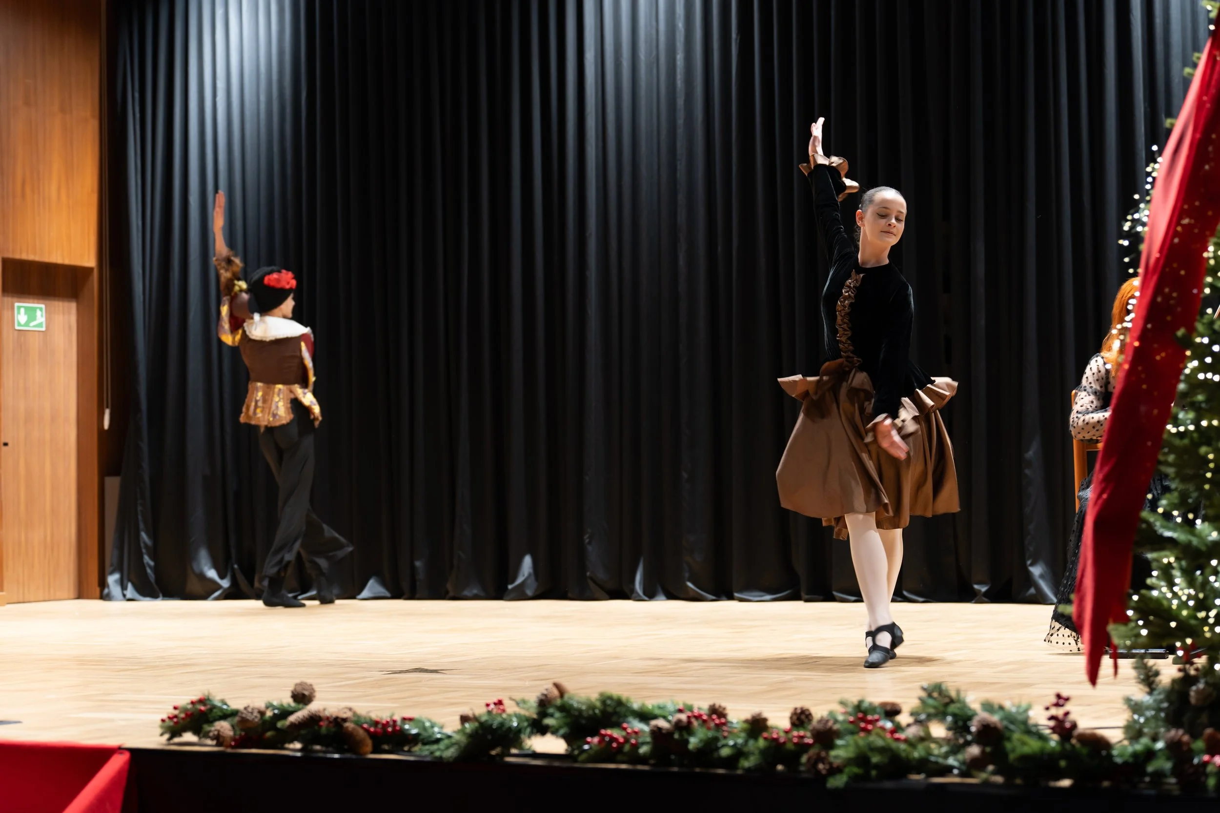 A ballet dancer performing on stage during a Christmas themed event, with children dressed in festive costumes and decorated Christmas trees in the background.
