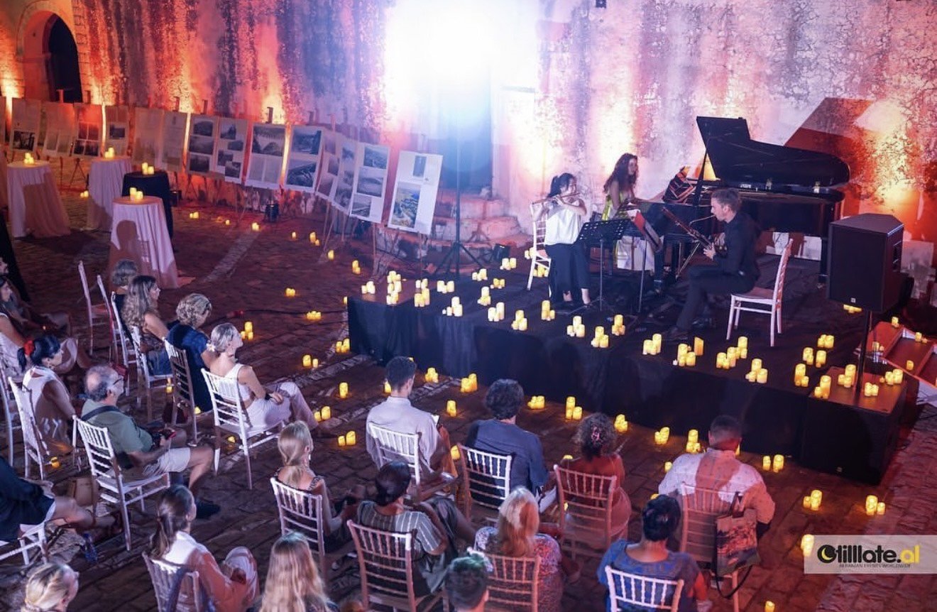 An in-door musical performance with an audience seated in chairs, surrounded by candle lights, with a trio of musicians playing on a stage, including a pianist, a violinist, and a flutist, in a large rustic hall with exposed brick walls.