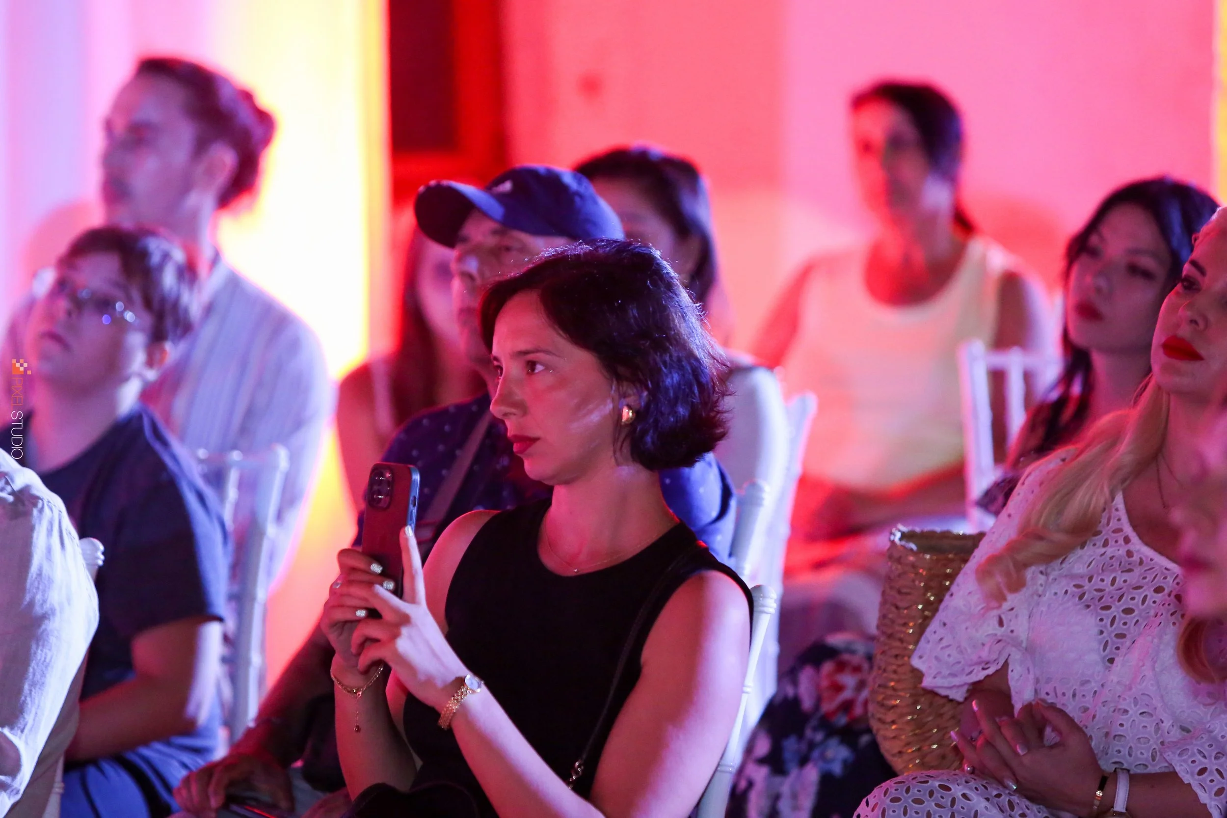 Group of women sitting in a theater or auditorium, some looking at their phones, others looking ahead, with colorful lighting in the background.
