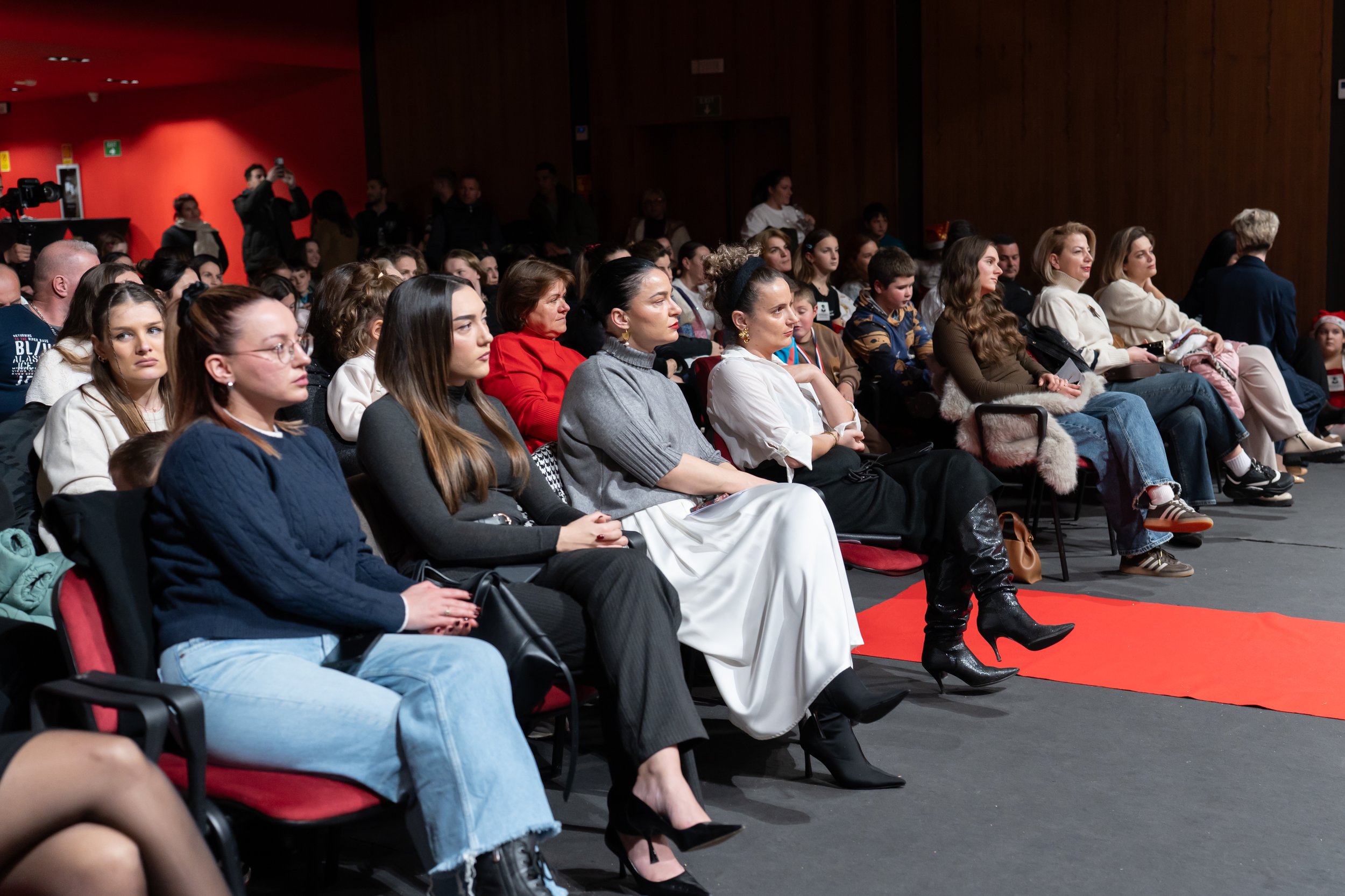Audience seated in chairs at an indoor event, some dressed casually and others more formally, with a few standing in the background against red and wooden walls.