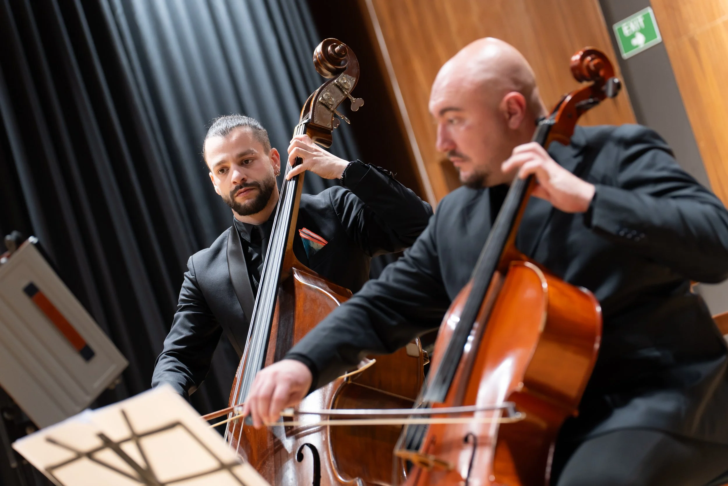 Two men playing string instruments, a woman with a beard and long hair tied back, and a bald man, performing on stage with music stands and sheet music, and a black curtain backdrop.