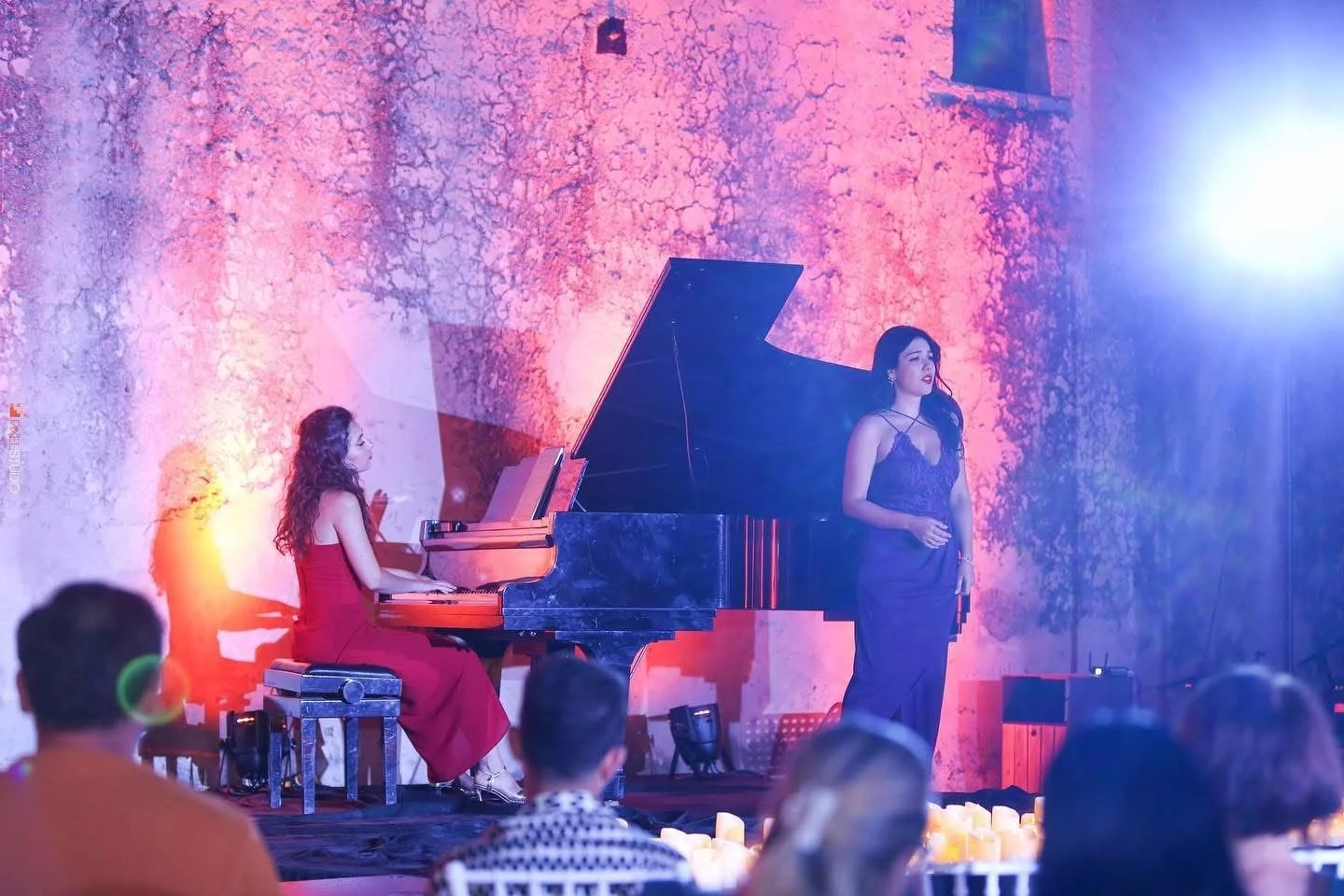 A woman in a black dress is singing while standing on stage in front of a pink and purple textured wall, with a pianist in a red dress playing a grand piano nearby, during a performance with audience members visible in the foreground.