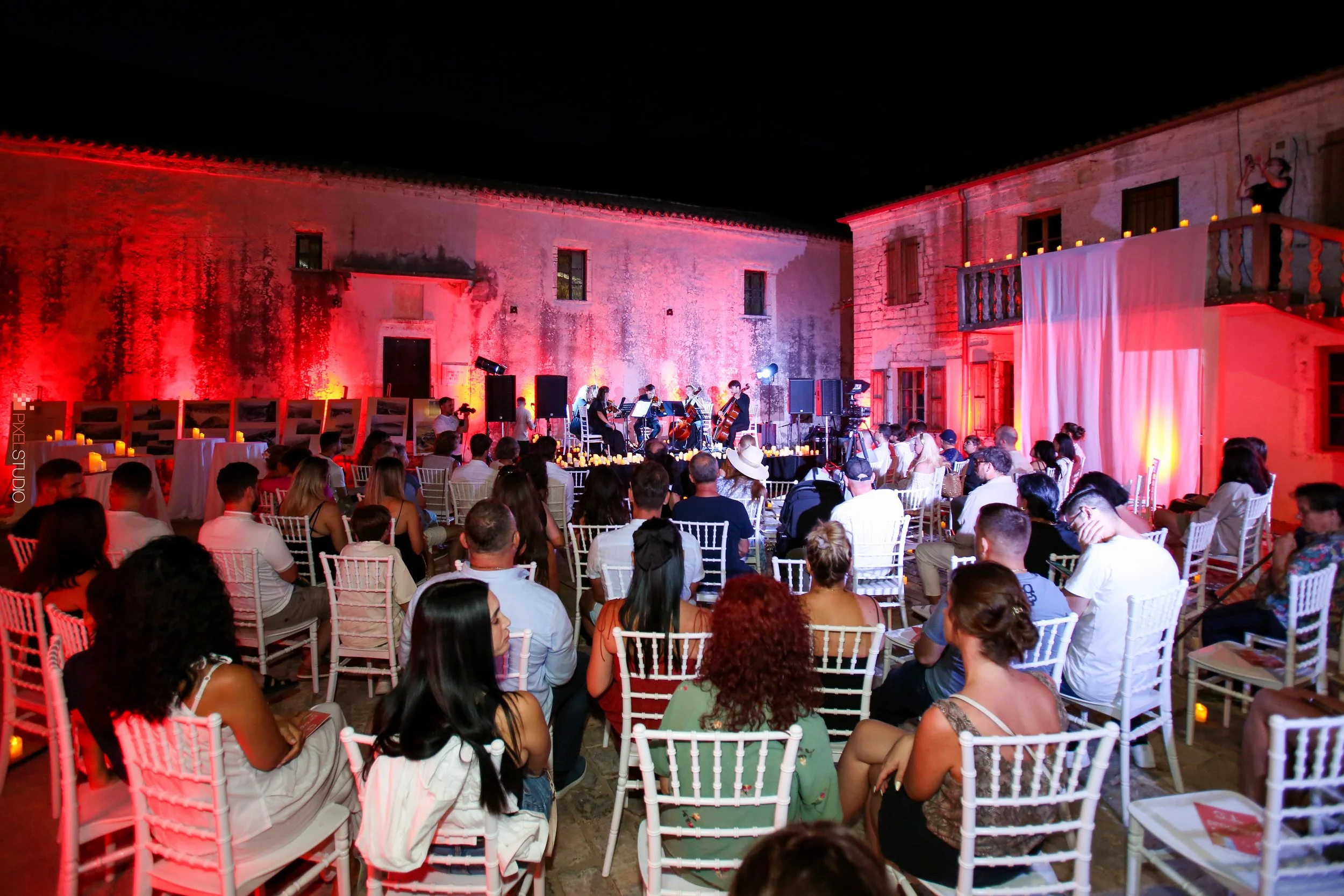 Outdoor concert venue at night with seated audience, musicians performing on stage, surrounded by old stone buildings and lit candles.