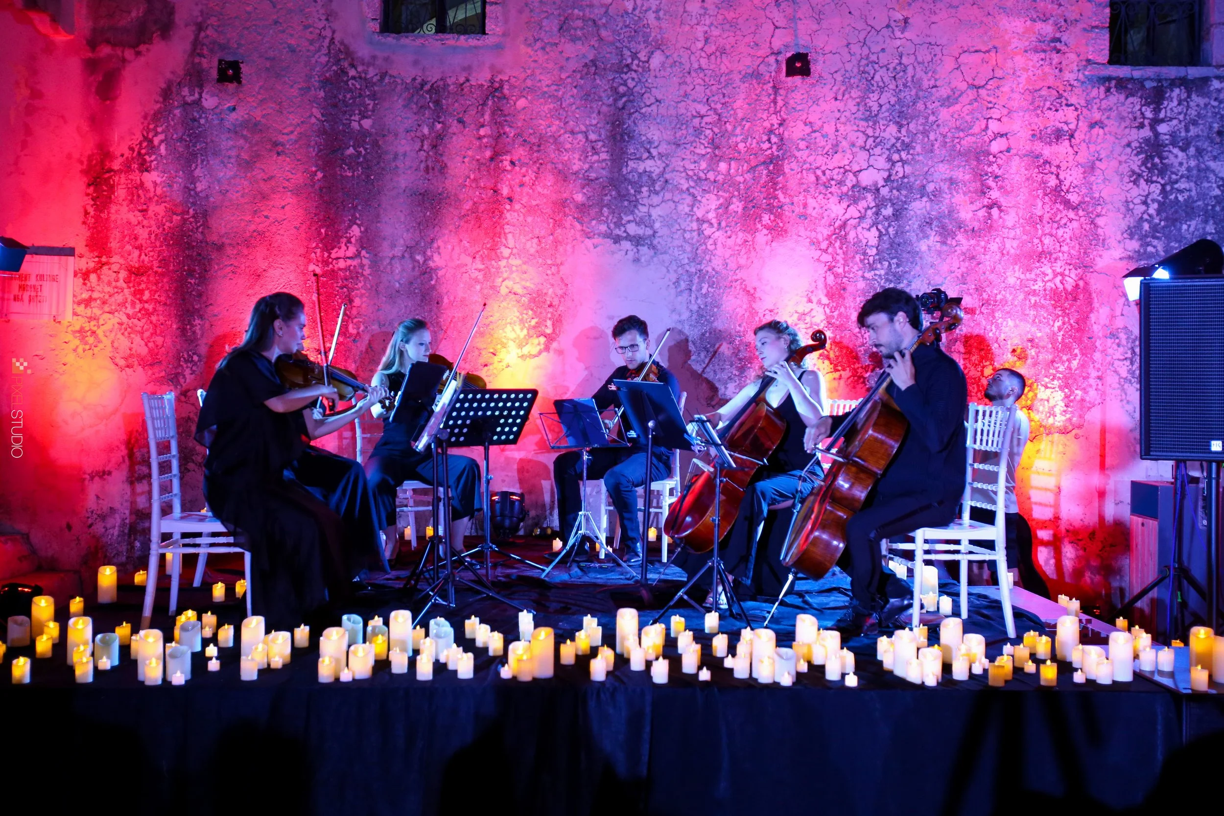 A group of musicians performing with string instruments on a stage decorated with candles and illuminated by colorful pink and purple lighting, with a rough textured wall in the background.