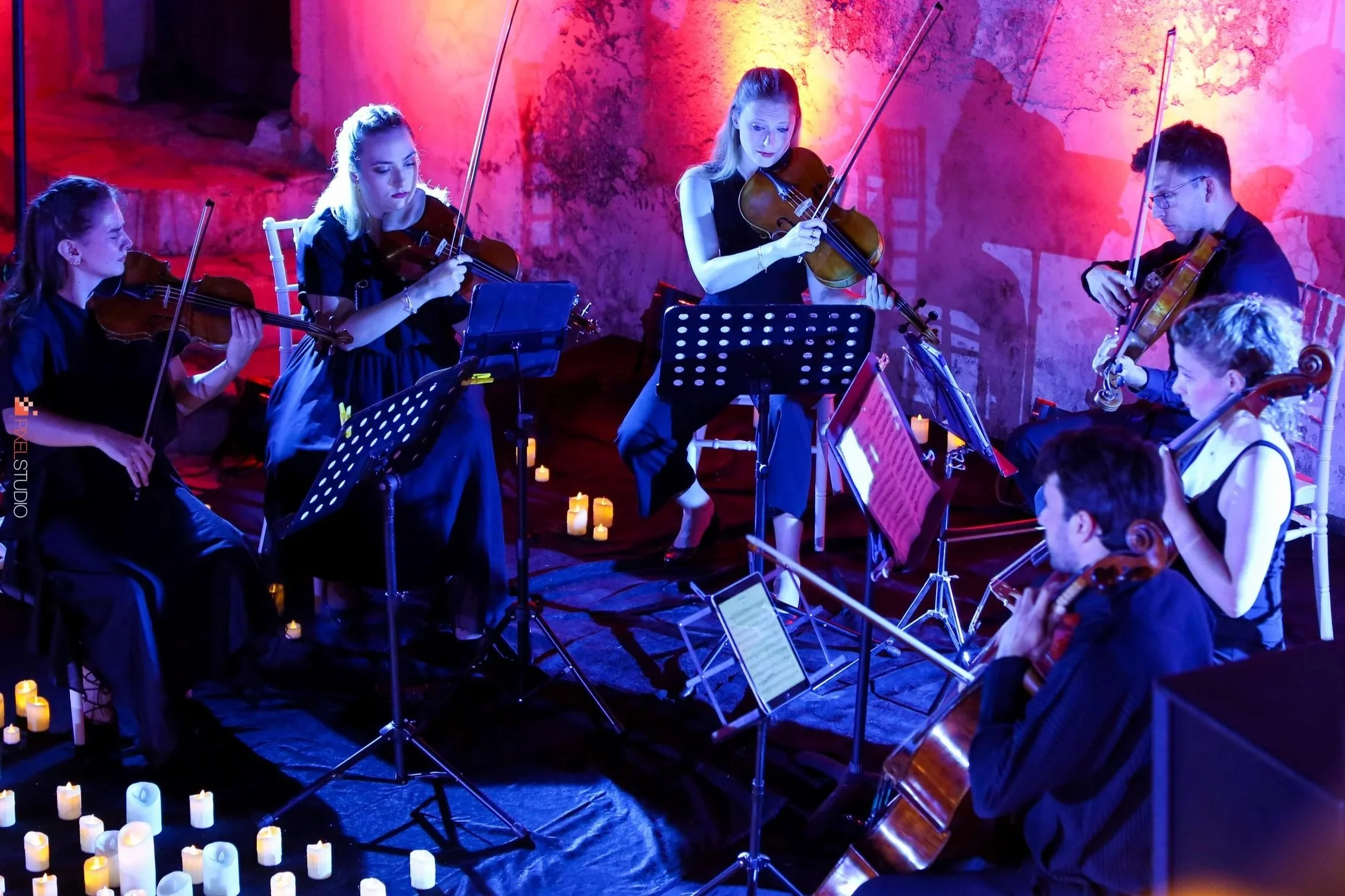 A group of six musicians playing violins and a cello during a concert with candlelights on the floor and colorful lighting on the wall.