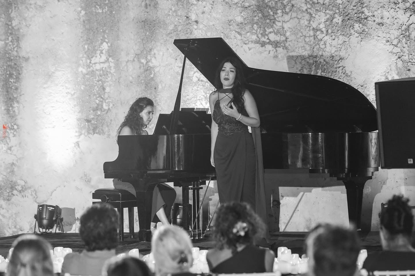 Two women performing a song on stage with a grand piano. One woman is singing while standing, and the other is playing the piano. The audience's heads are visible in the foreground.