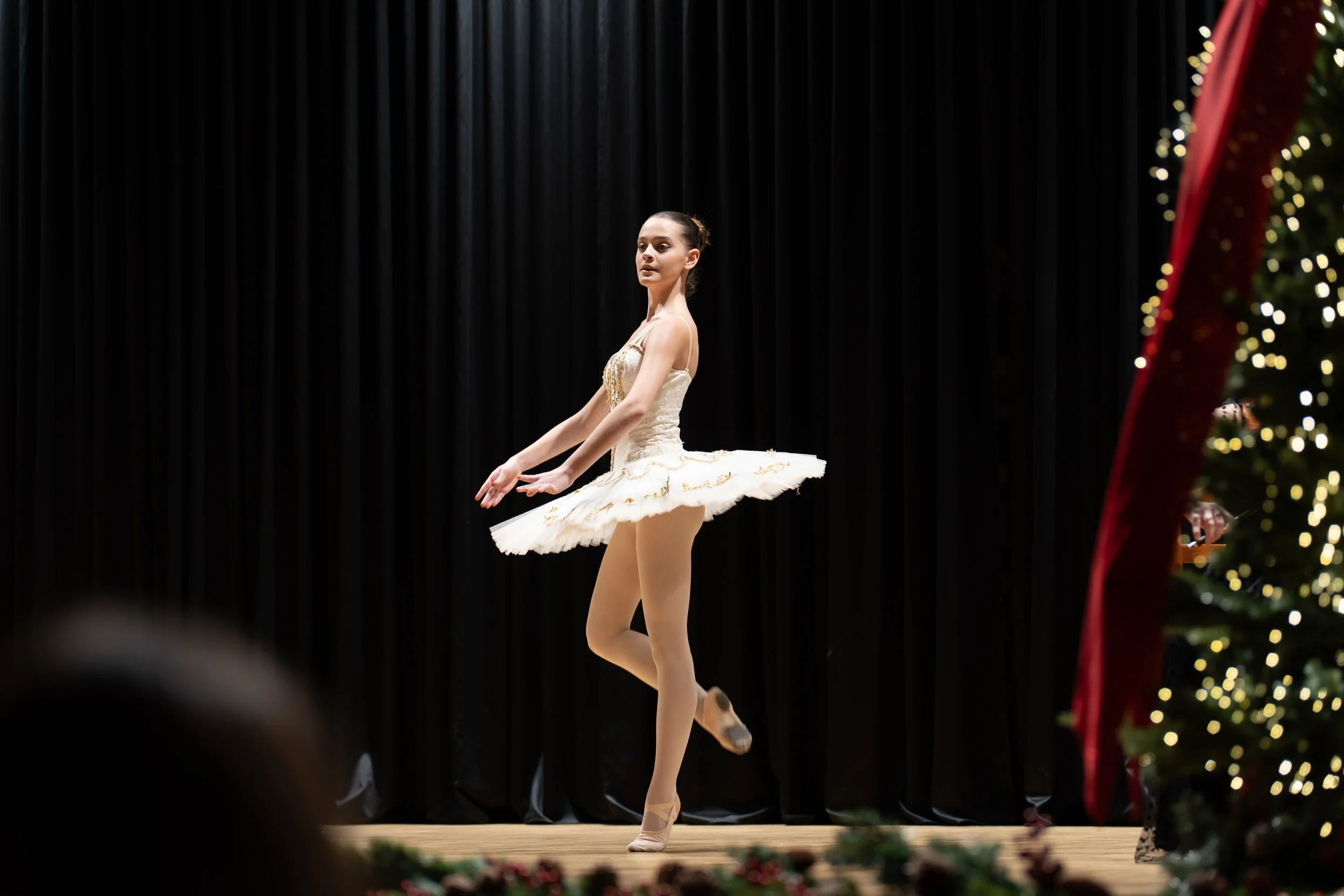 Ballet dancer in a white tutu practicing on stage with black curtains behind and holiday decorations in the foreground.