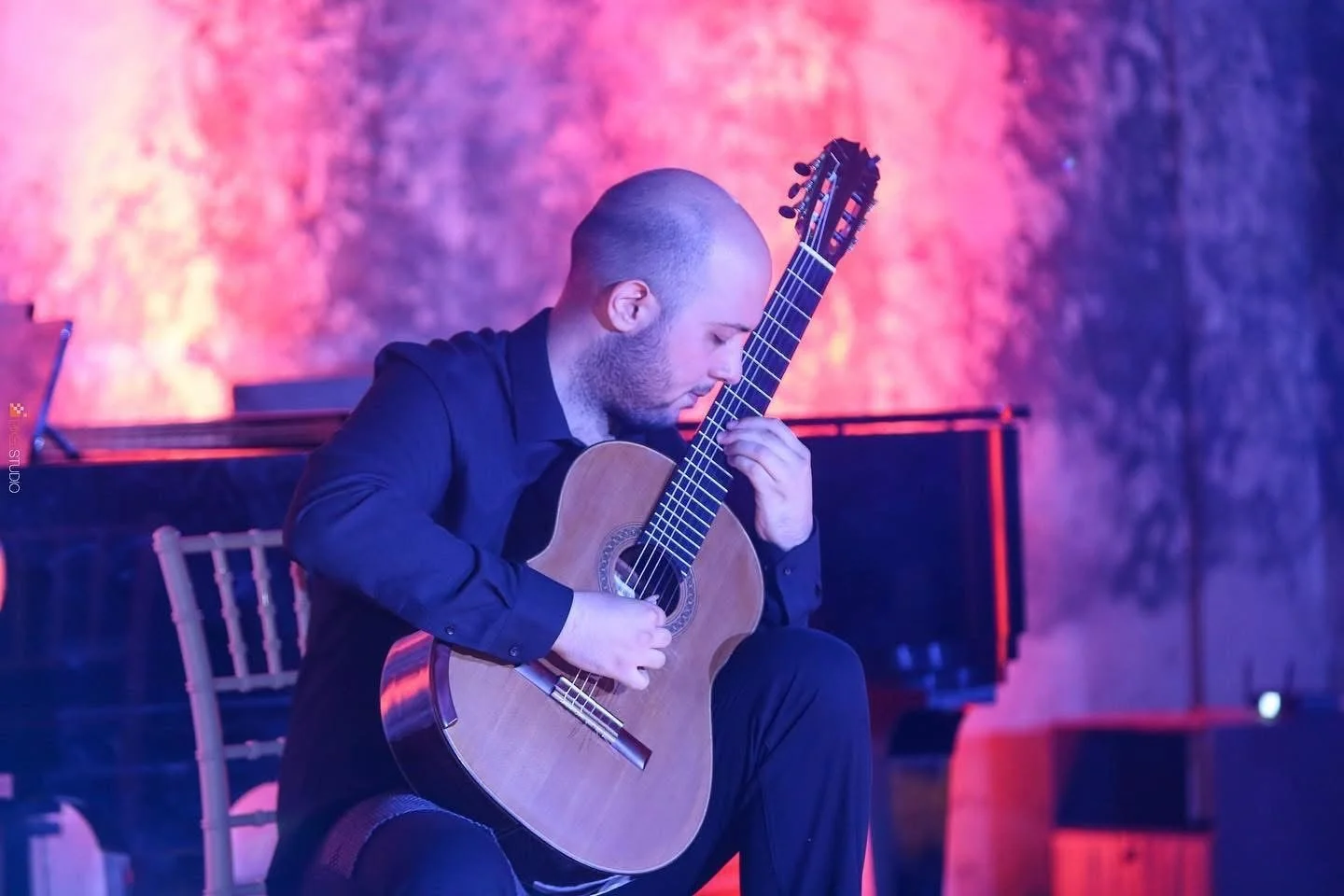 A man playing an acoustic guitar on stage in a dimly lit performance space with a colorful, textured backdrop.