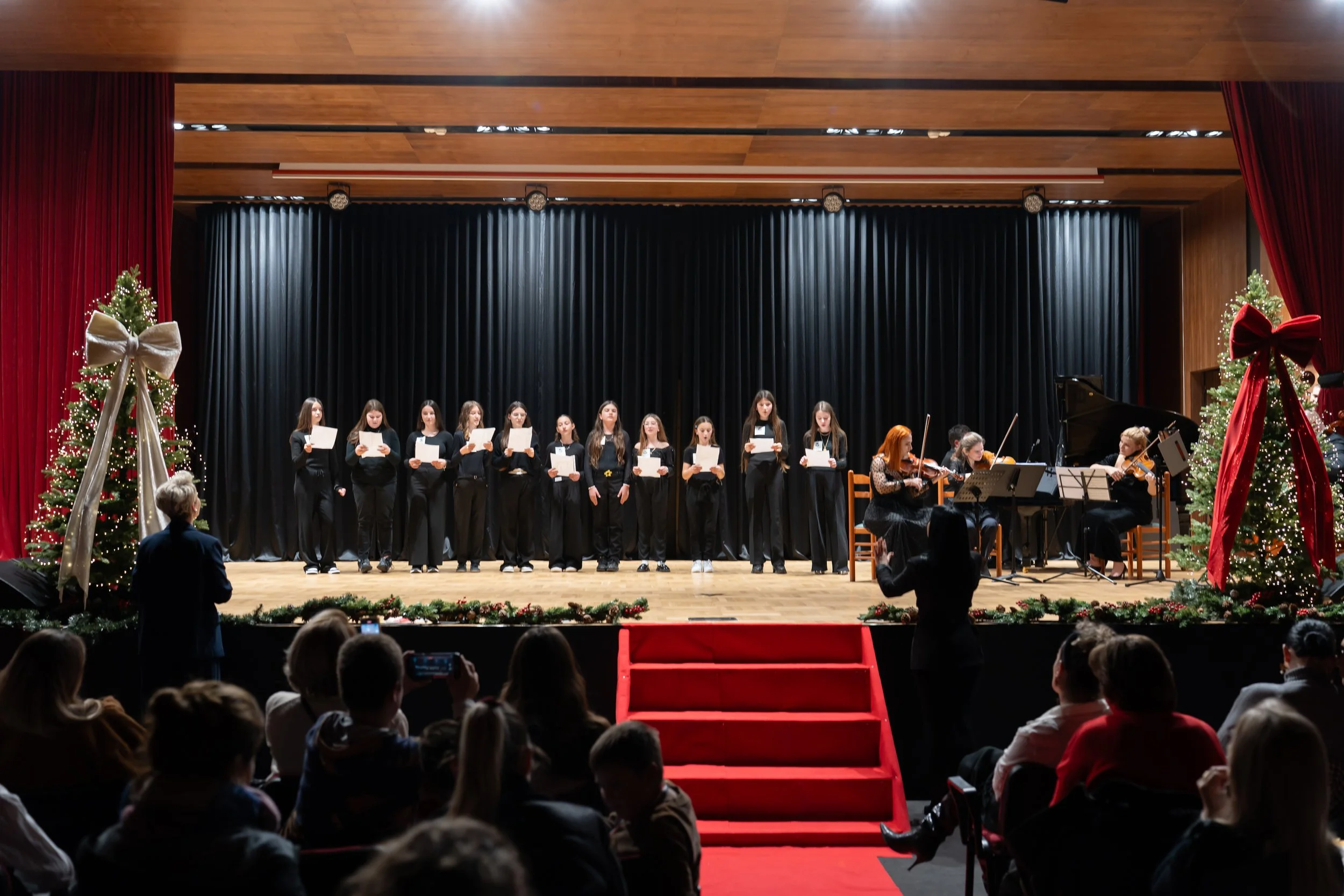 A choir of young women and two women playing violins are performing on stage decorated with Christmas trees and holiday ornaments, with an audience watching.