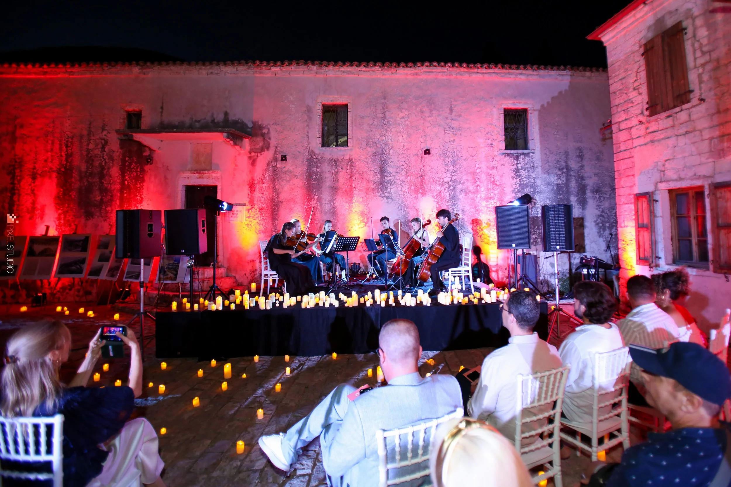 An outdoor musical performance at night with a string quartet on stage illuminated by pink and yellow lighting. The stage is decorated with numerous candles, and there is a rustic building wall in the background. Audience members are seated and some are taking photos.