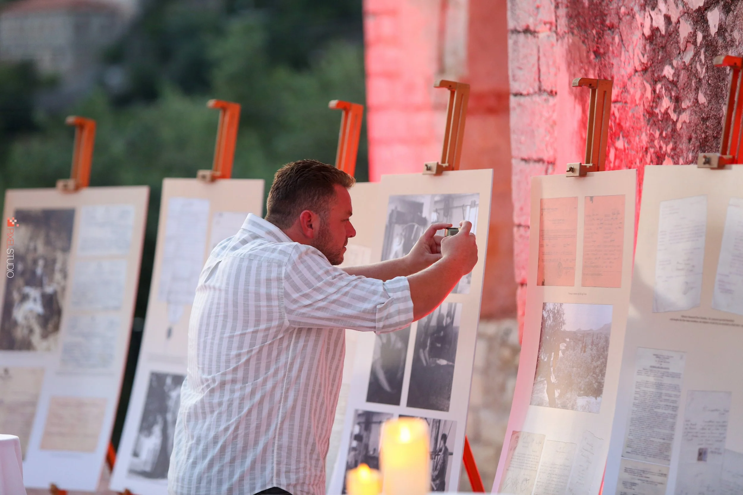 A man in a white checkered shirt is taking photos of display boards with photographs and documents during an outdoor event, with a brick wall and blurred greenery in the background.