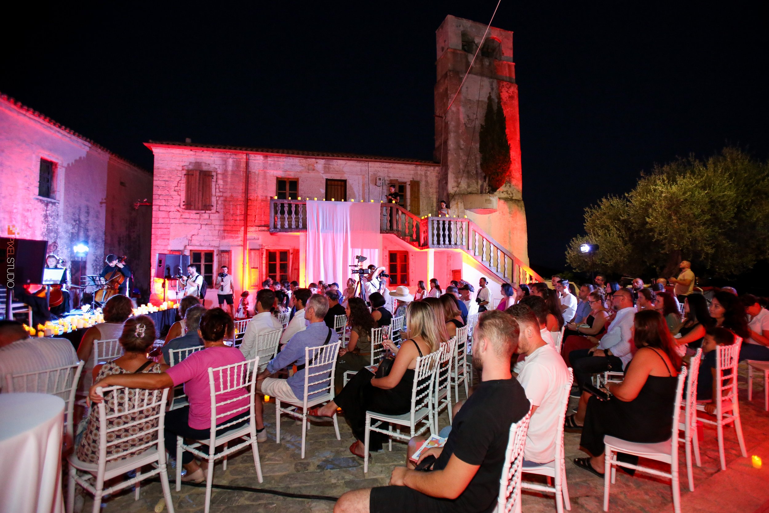 Nighttime outdoor gathering with many people seated on white chairs, a stage with musicians performing, and a historic stone building illuminated with red lights.