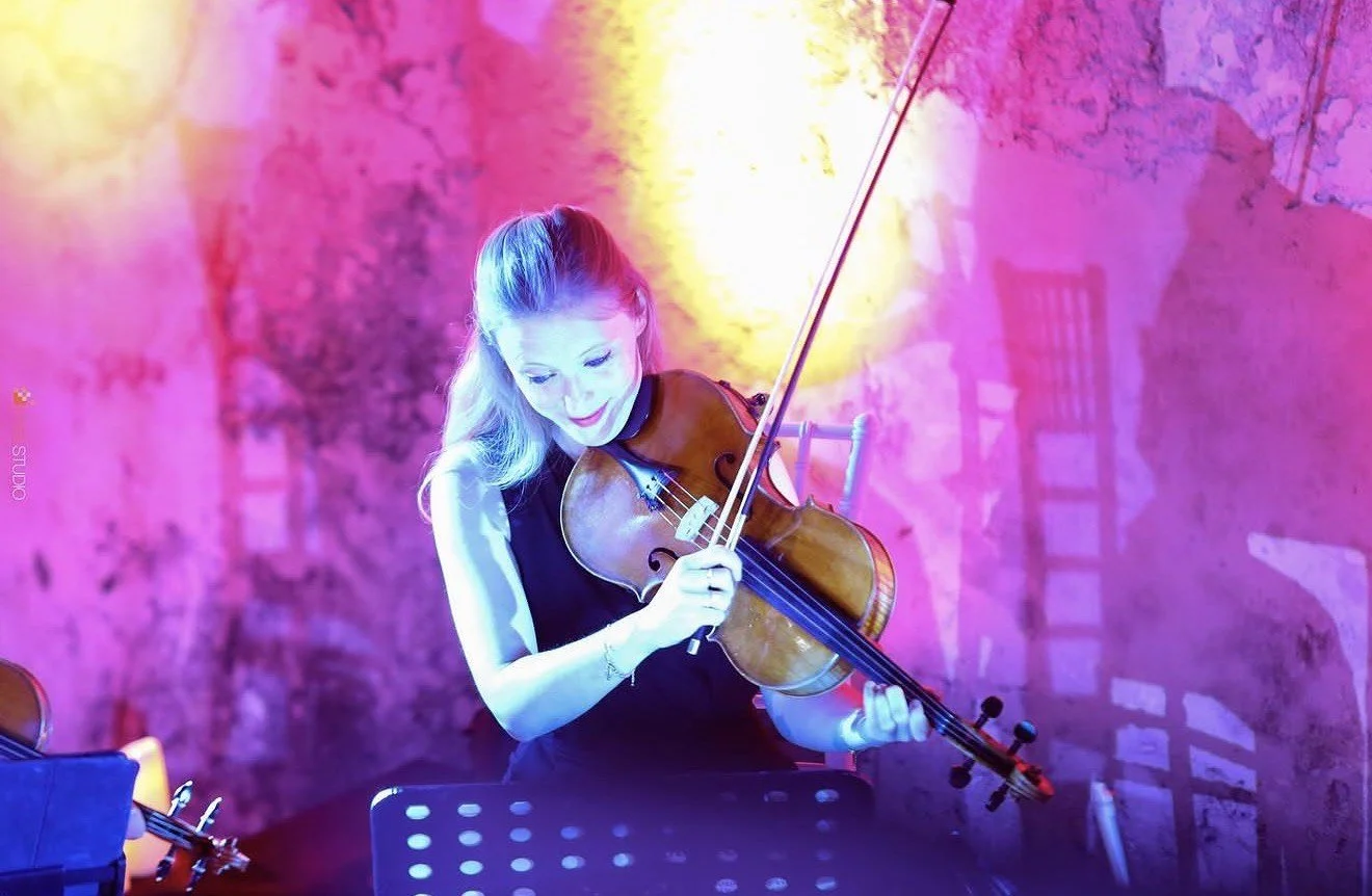 A woman playing the violin in a colorful, vibrant setting with pink, purple, and yellow lighting and a textured background.