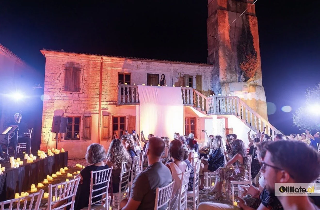 An outdoor night event at a historic stone building illuminated with pink lights, with guests seated facing a stage with candles, and candles on tables, under a dark sky.